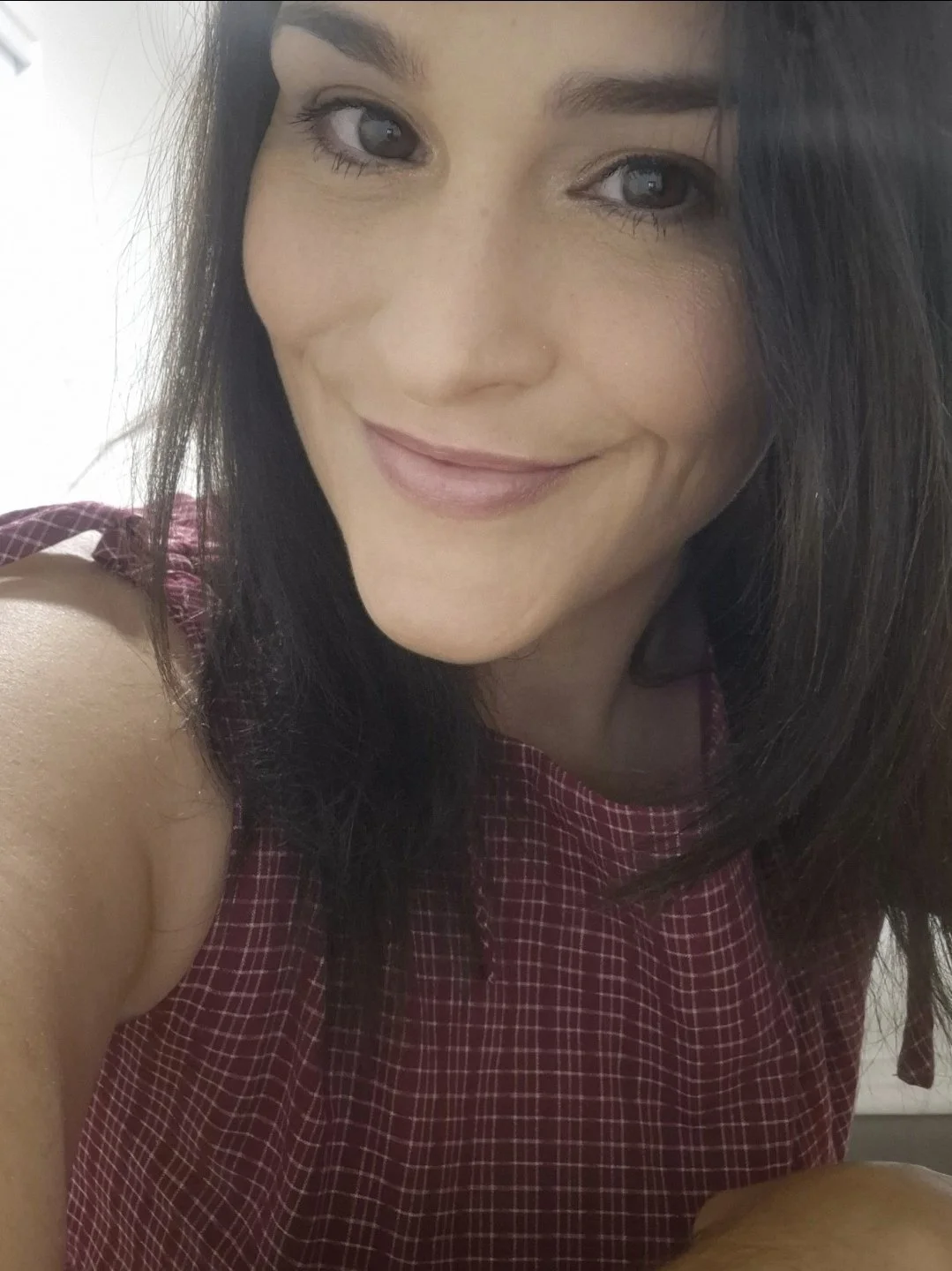 Close-up photo of a woman with long dark hair, smiling, wearing a sleeveless red checkered top.