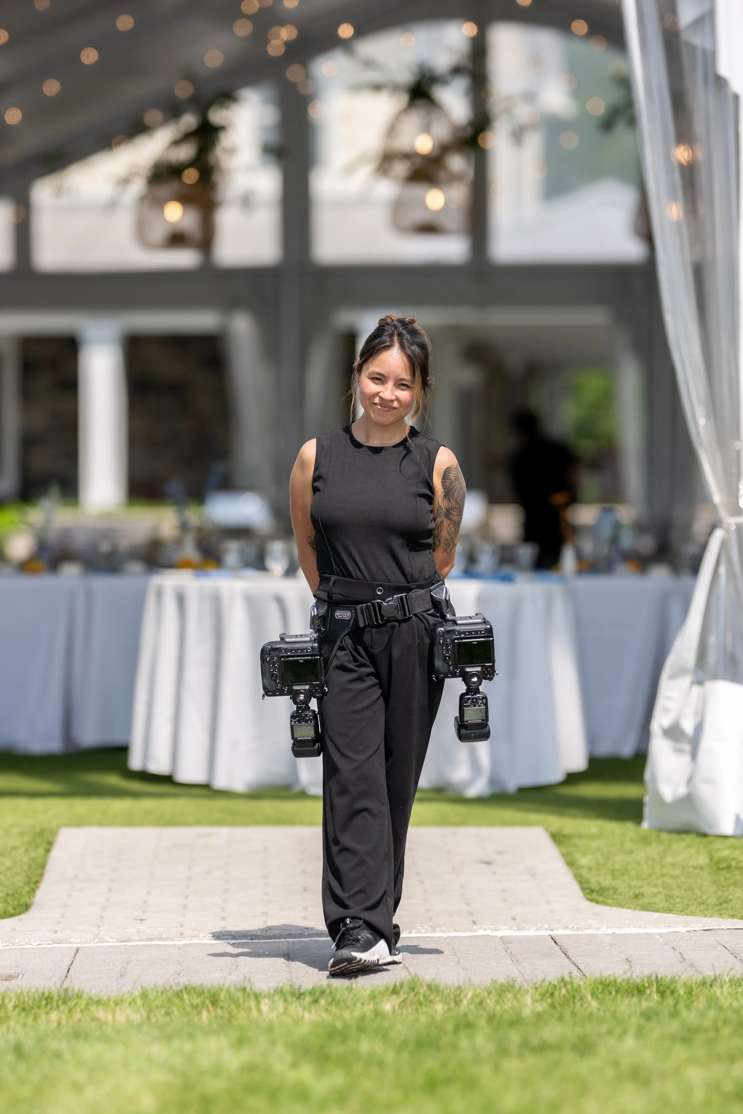 A woman in black athletic clothing walks on a pathway in a garden with a tent in the background. The tent has string lights and potted plants hanging from it.