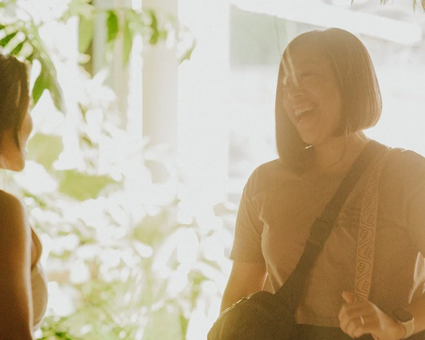 Two women smiling and engaging in conversation indoors with sunlight and greenery in the background.