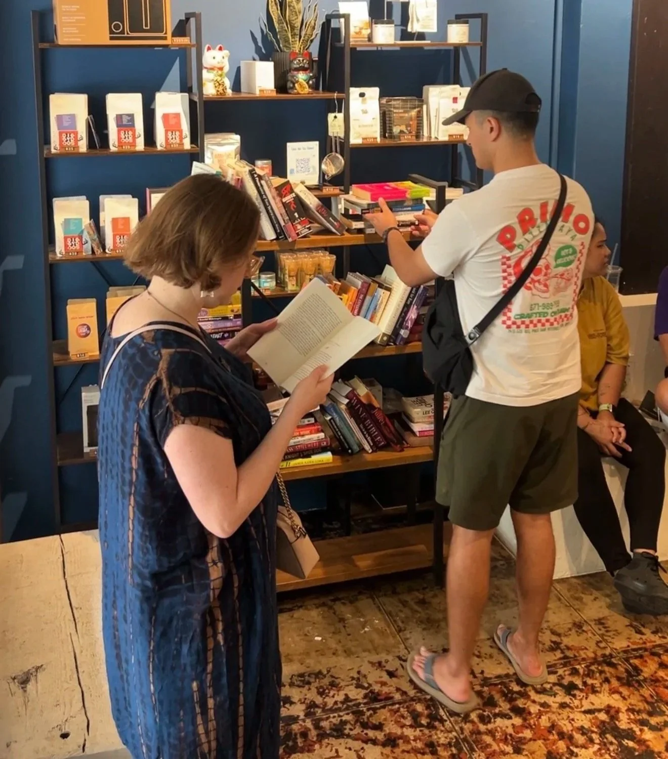 People browsing books at a bookstore or library. One person is reading a book, another is looking at books on a shelf, and there are others seated nearby.