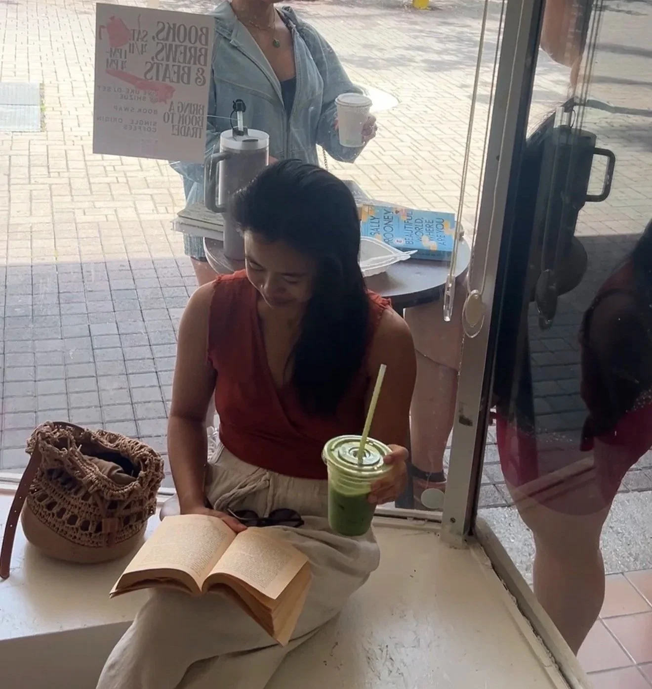Woman sitting indoors with an open book, a wicker purse, and sunglasses on her lap, holding a green smoothie with a straw. Outside through the glass, two women are visible, one holding a coffee cup and the other with a flyer on the table behind the g
