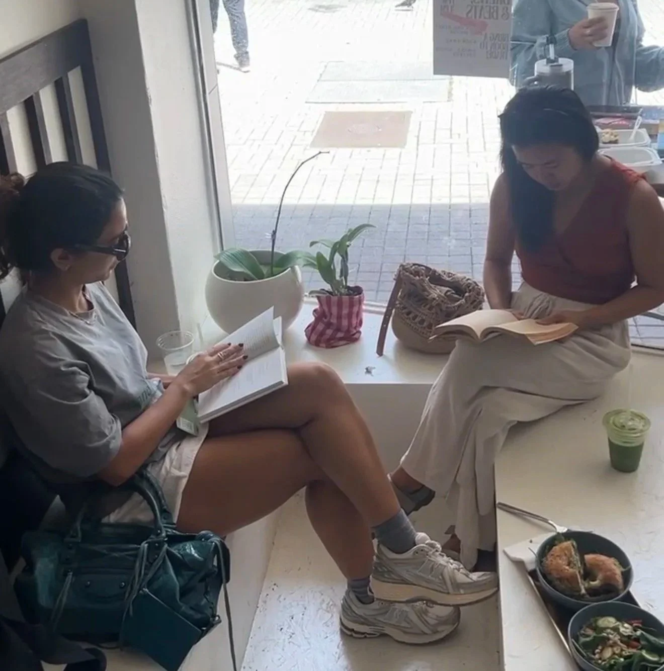 Two women sitting inside a cafe, reading books. One woman with dark hair and glasses is seated on a chair, wearing a gray t-shirt, shorts, and sneakers, with a black bag at her feet. The other woman with dark hair is standing, wearing a red top and b