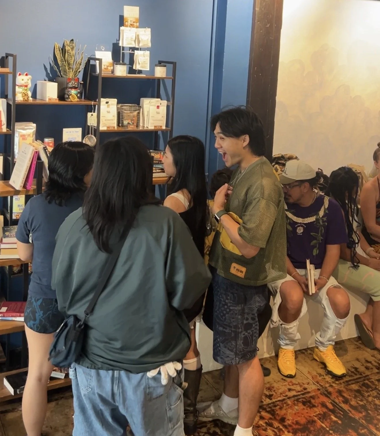 A group of people are chatting and sitting in a cozy bookstore or café. Several individuals are standing and engaged in conversation, while others are seated, holding books. The space is decorated with blue walls, wooden flooring, and shelves filled 