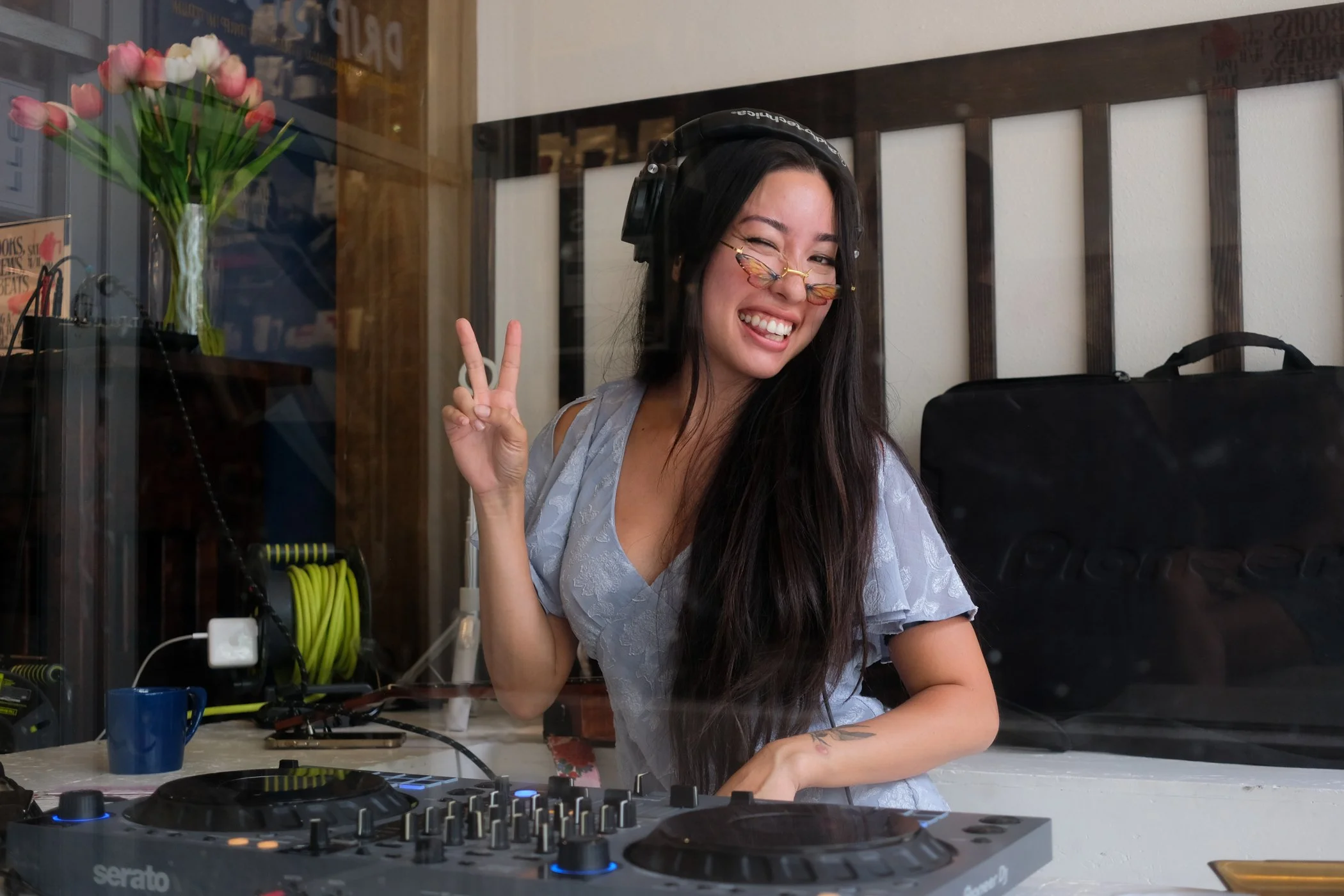 A young woman with long dark hair, wearing butterfly-shaped glasses, making a peace sign with her right hand, smiling, and standing behind DJ equipment in a room with a glass window, a blue mug, and a black bag.