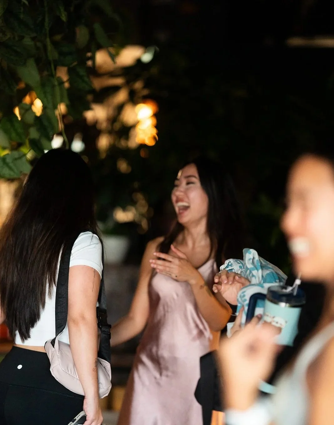 Three women are seen laughing and talking outdoors at night, with a blurred background of foliage and lights. One woman in the center is touching her chest with a joyful expression, while another on the right is holding a water bottle and wearing a patterned scarf. The third woman on the left has long dark hair and a backpack.