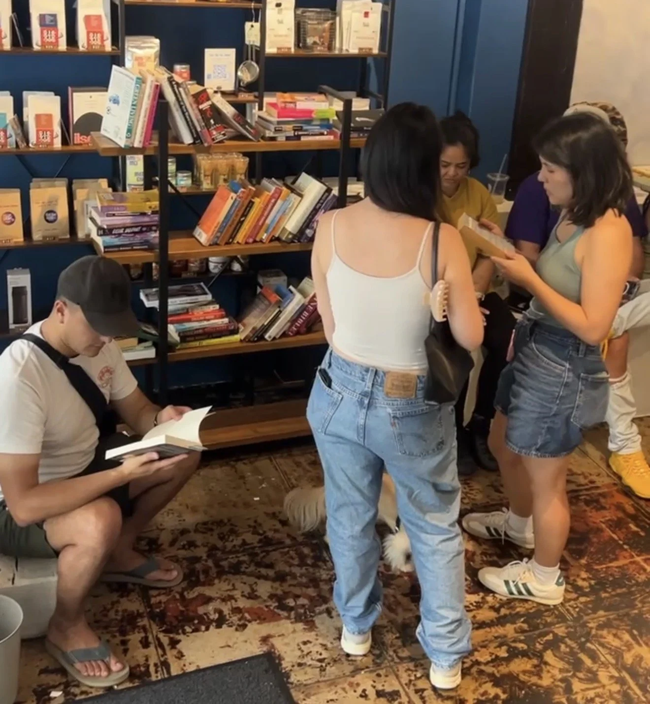Group of young women and a young man in a bookstore, with others looking at books on shelves and one person reading while sitting on the floor.