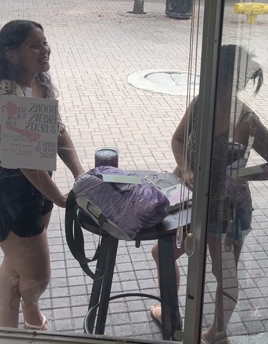 Two women are outside looking into a storefront window. One woman is smiling and holding a sign, while the other woman is leaning on a table with bags and glasses on it.