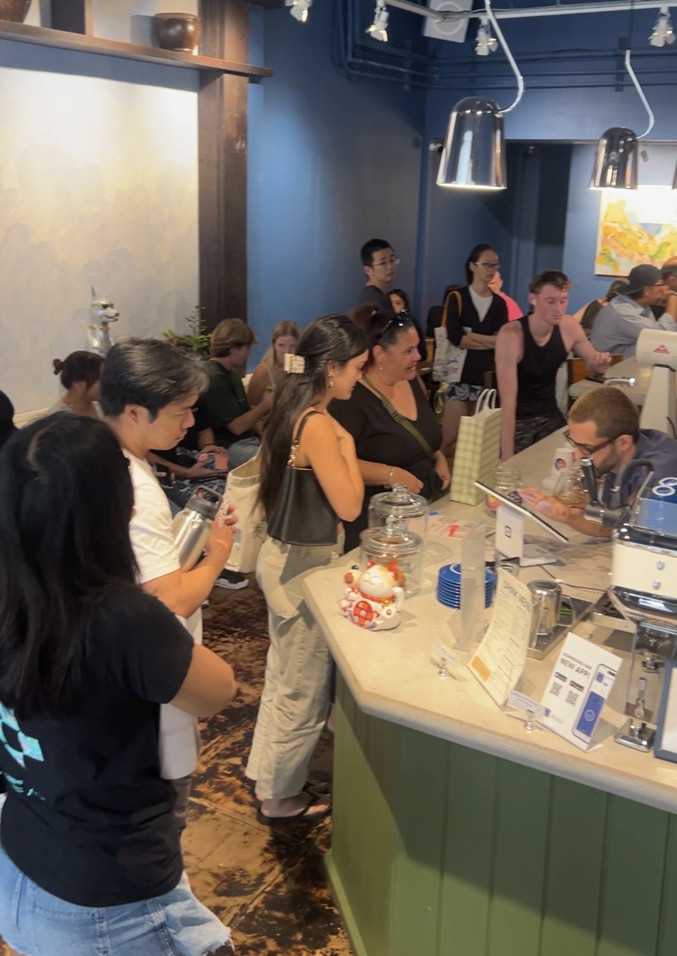 People standing in line at a café or bakery counter with menu items and promotional signs on display.