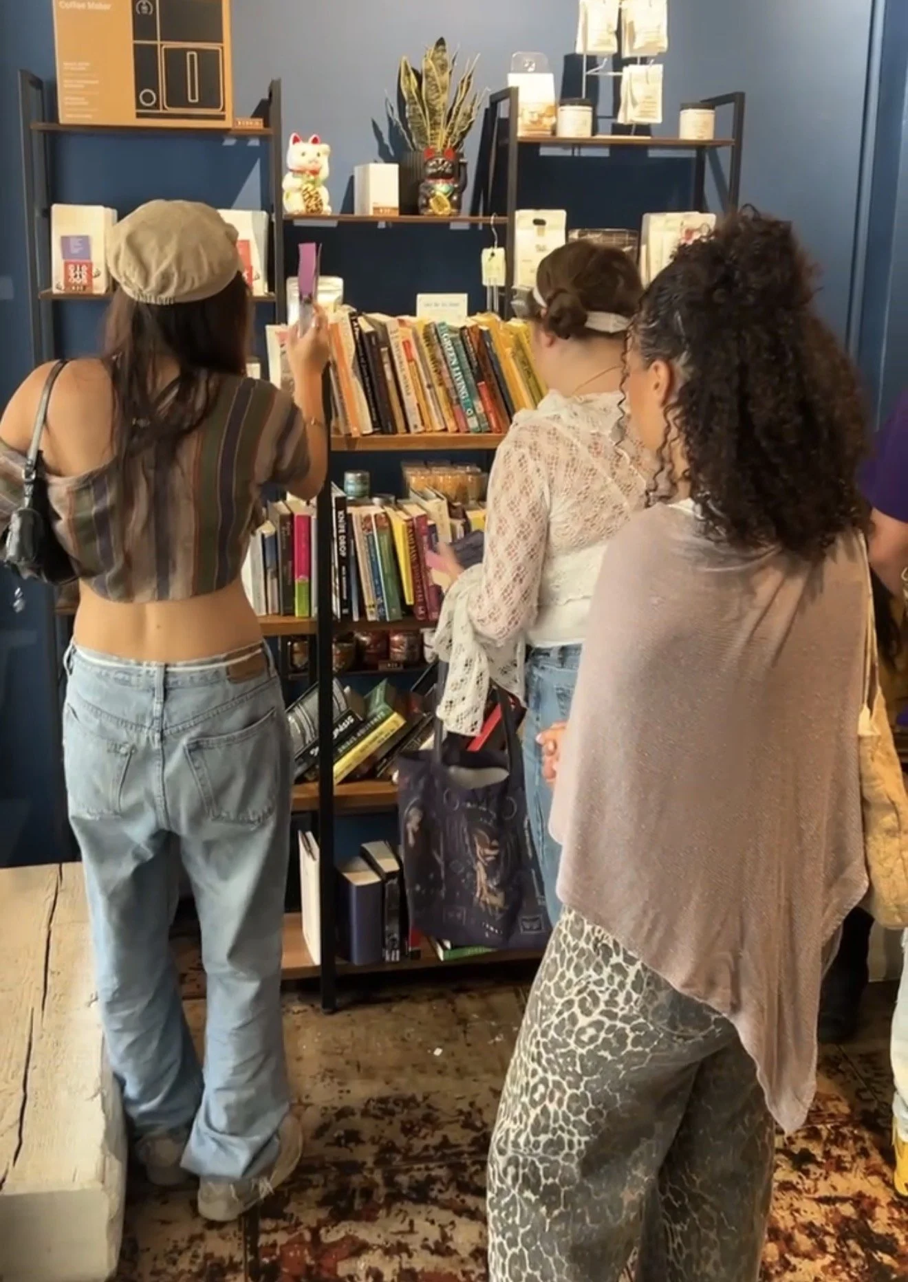 Three women browsing books at a bookstore or library. One woman is wearing a beige beret and loose jeans, looking at a book. The other two women are facing the shelves, one with curly hair and a light pink shawl, the other with either a bunt or updo 