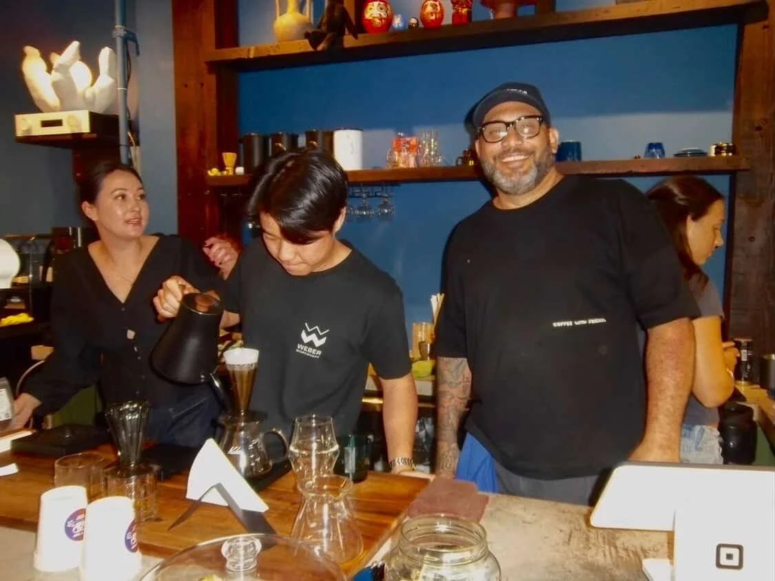 Barista pouring coffee at a busy coffee shop, with smiling man and women in the background.