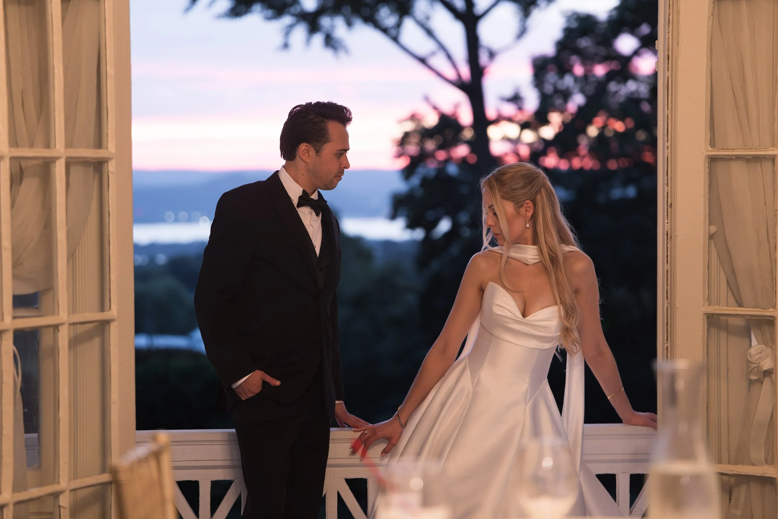 A man in a tuxedo and a woman in a white wedding dress stand close to each other at a window during sunset, holding hands, with a scenic landscape and a large tree in the background.
