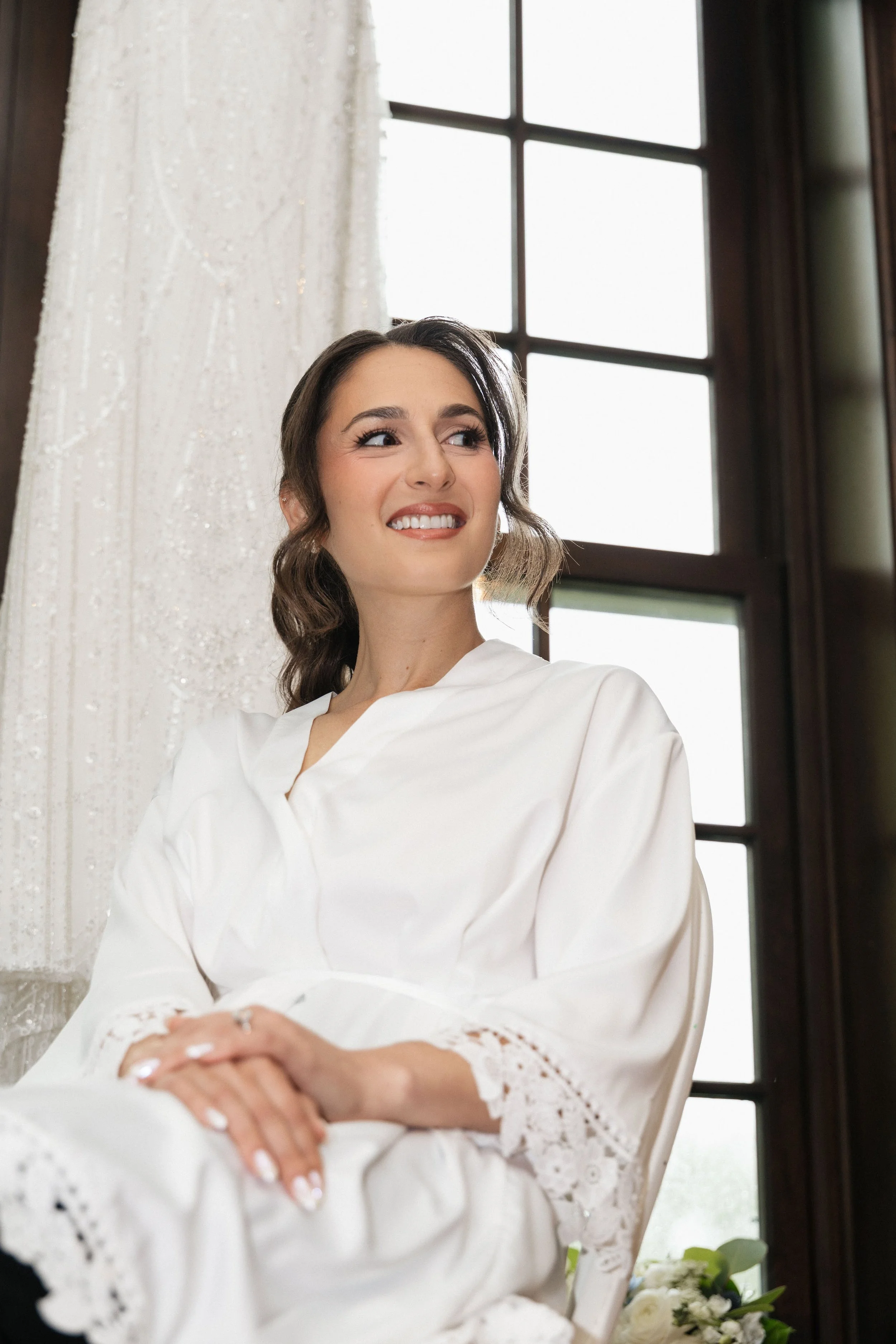 A woman with dark brown hair styled in loose waves, wearing a white satin robe with lace trim, smiling softly as she looks to her left. She is seated indoors near a window with dark wood framing and sheer white curtains, with a bouquet of white flowe
