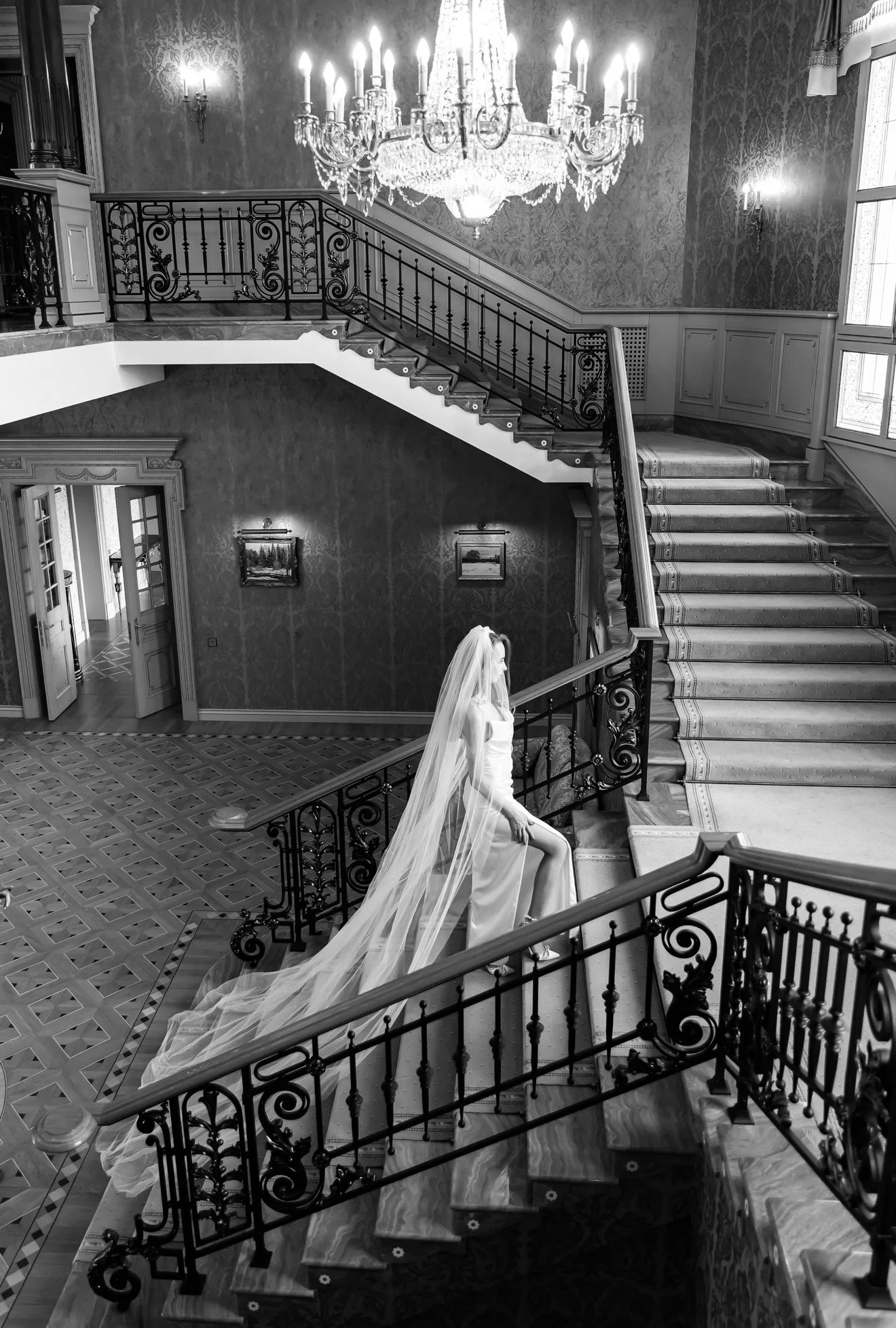 A woman in a wedding dress with a long veil sitting on the stair railing of an elegant, chandelier-lit staircase in a richly decorated interior. The space features ornate walls, framed artwork, and large windows.