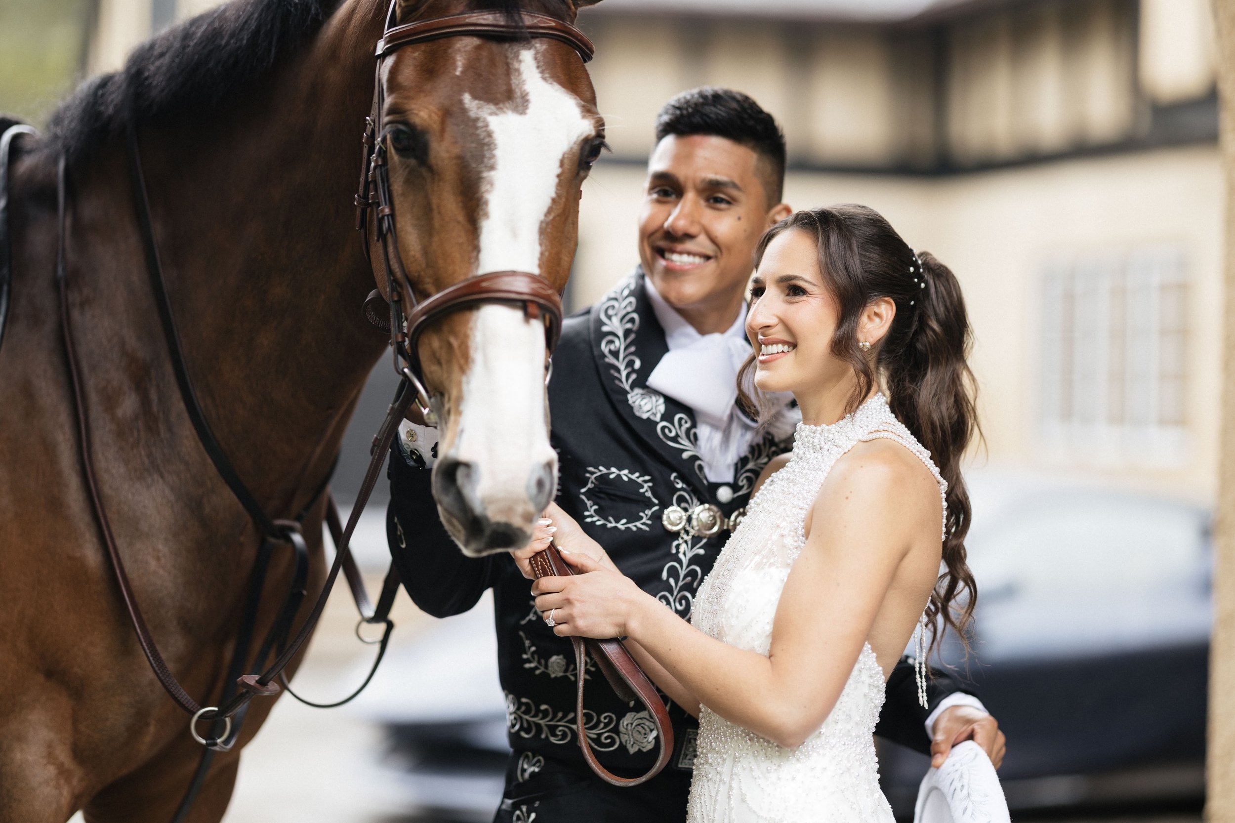 A bride and groom smiling and holding hands while standing next to a brown horse with a white blaze on its face, in a stable or barn.