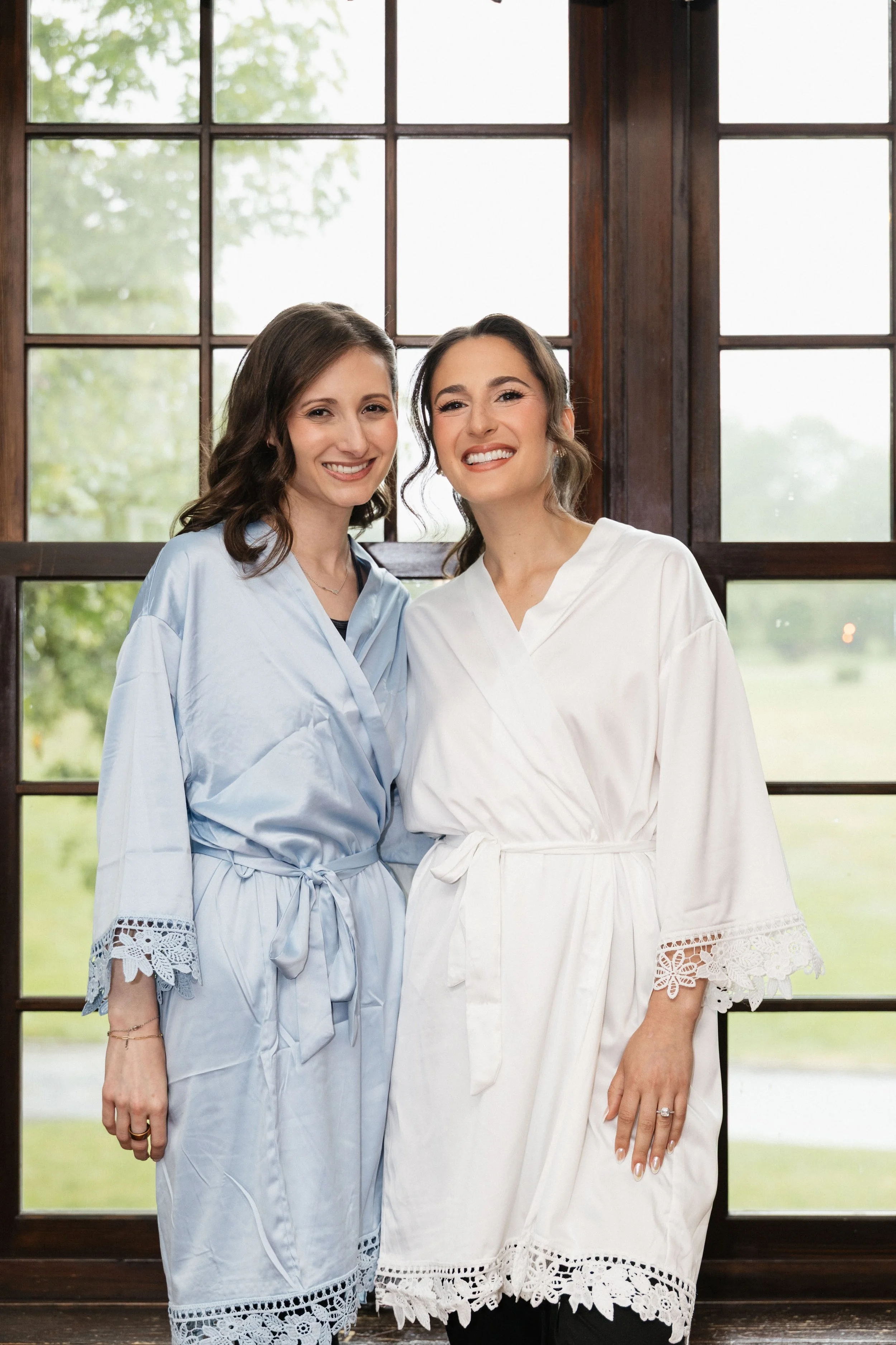 Two women standing close together inside in front of large windows with green scenery outside. One woman is wearing a light blue dress with lace details, and the other is in a white dress with similar lace details on the sleeves and hem. They are smi