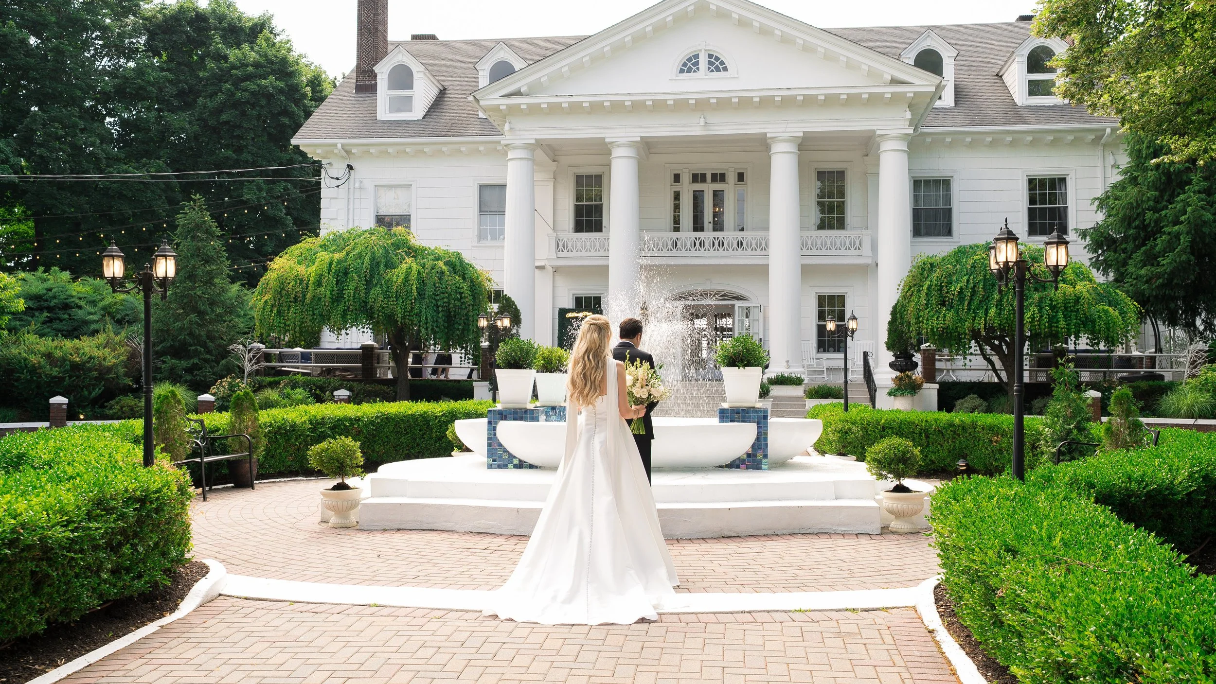 A bride in a white wedding gown standing in front of a fountain outdoors, near a large white house with a balcony, surrounded by greenery and street lamps.