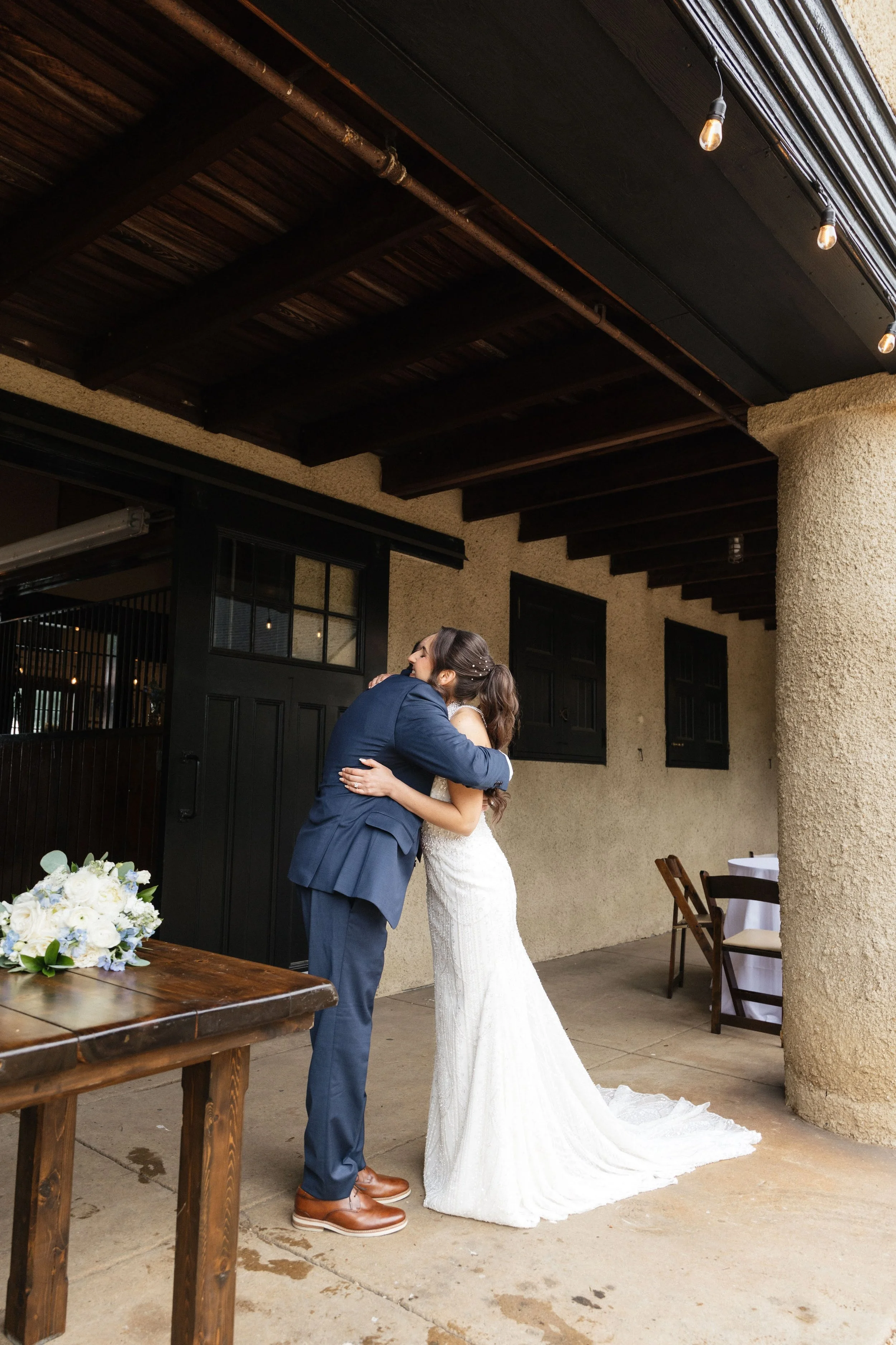 A couple, dressed in wedding attire, hugging on a covered outdoor patio with string lights and rustic decor.