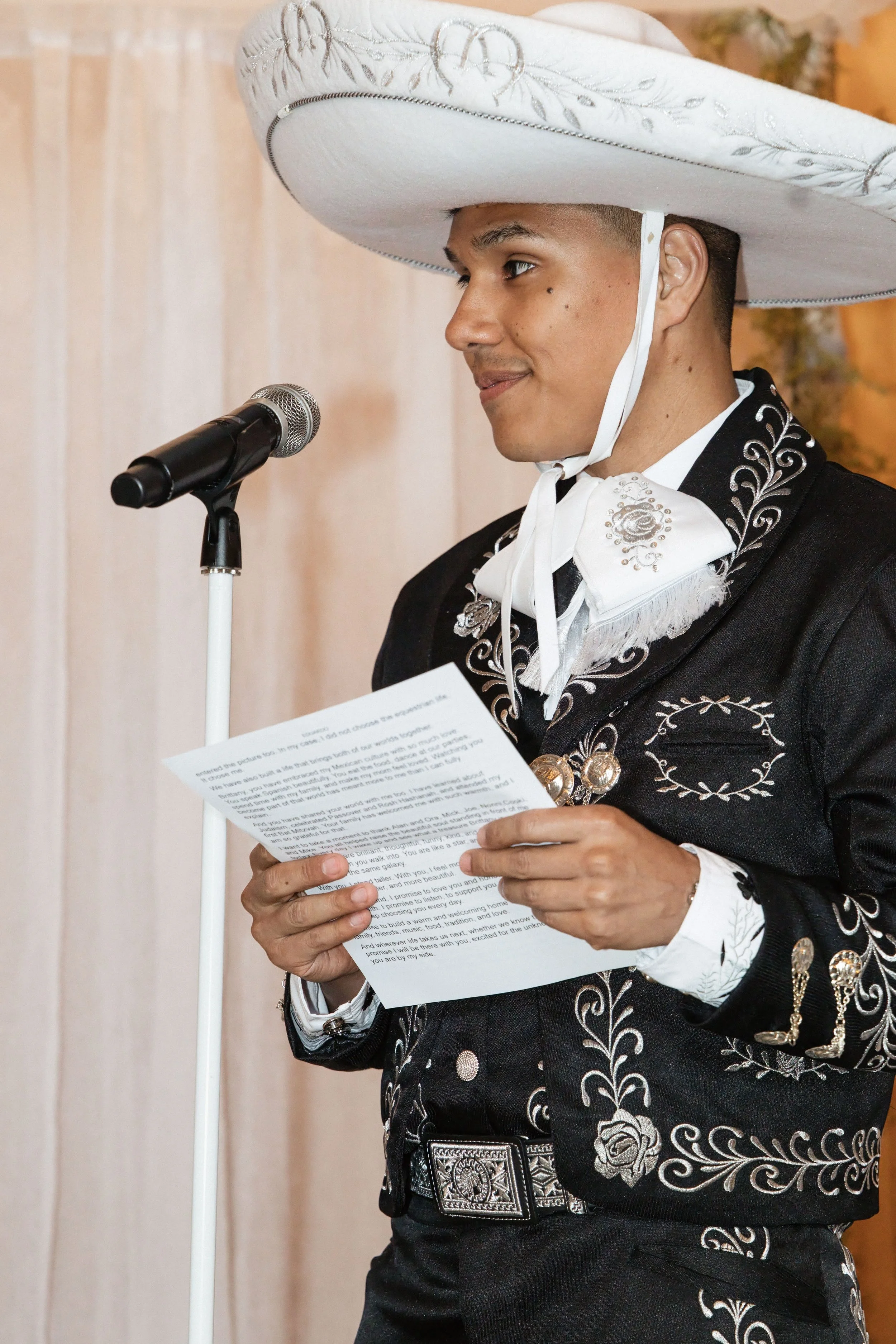 A man dressed in traditional Mexican charro attire is speaking into a microphone while holding a piece of paper. He is wearing a large white sombrero with embroidered silver designs, a black suit with intricate white embroidery, and a large belt buck