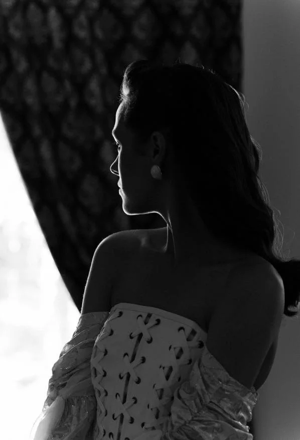 A woman with long hair looking out a window, silhouette style, black and white photo, sitting near curtains.