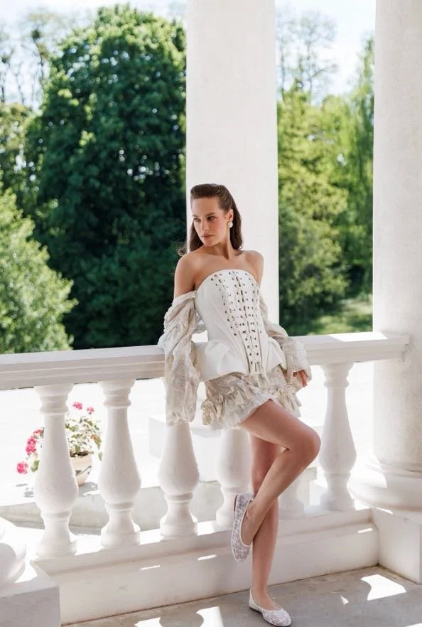 A woman in a white off-shoulder dress with lace details and ruffled hem, standing on a balcony with white balustrades and columns, outdoors with green trees in the background.