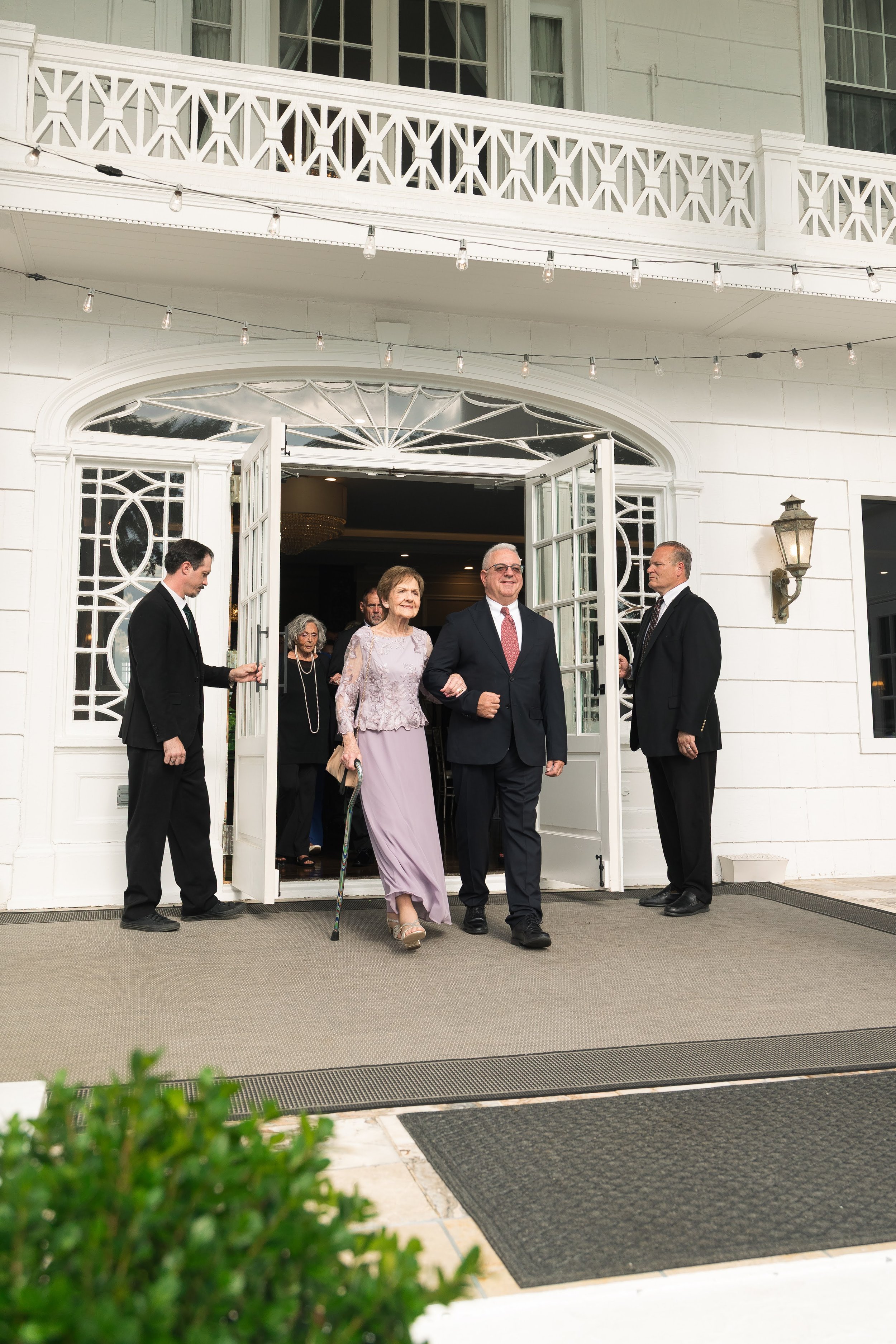A group of people, including an elderly woman with a cane, and a man in a suit, exiting a white building with open double doors, while others stand nearby. The building has a decorative balcony and hanging string lights.