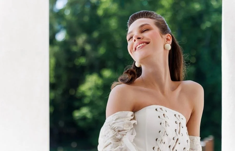 A young woman smiling outdoors, wearing a strapless white top and large pearl earrings, with green trees in the background.