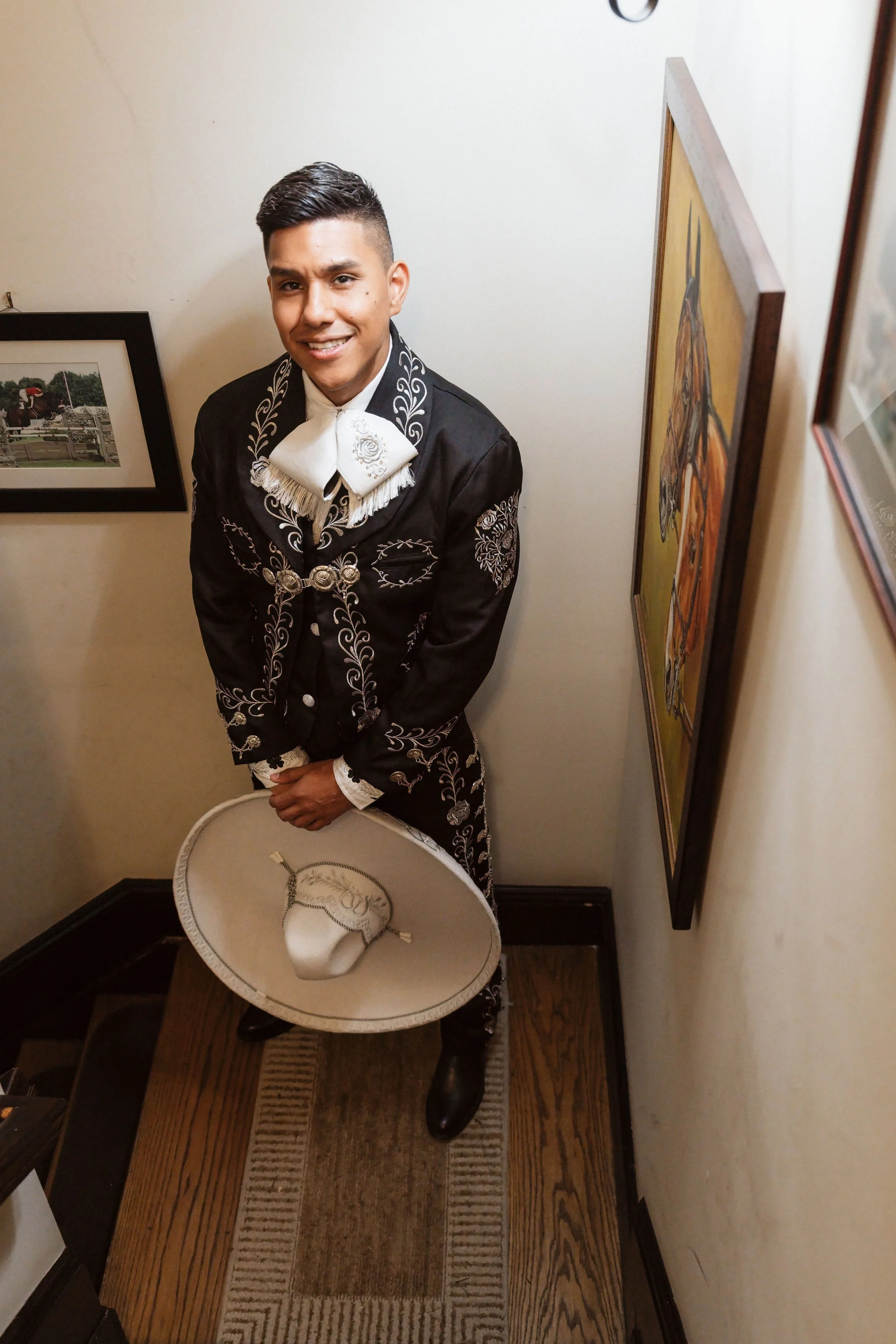 Young man dressed in traditional Mexican charro attire, wearing a black embroidered suit and white bow tie, standing in a corner of a room with framed artwork on the walls, holding a white sombrero on a small side table.