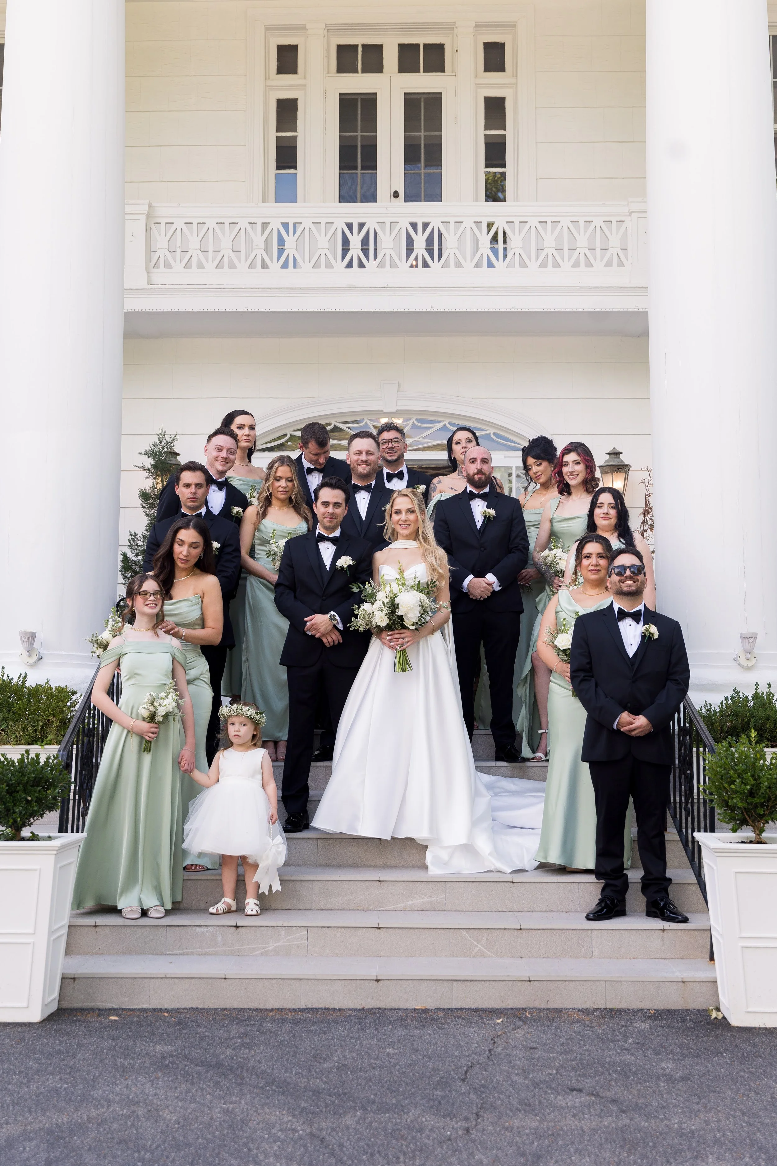Group of wedding guests, including adults and children, standing on the stairs of a white house with large white columns, celebrating a wedding. The bride is at the center holding a bouquet, and the groom is beside her. The guests are dressed in form