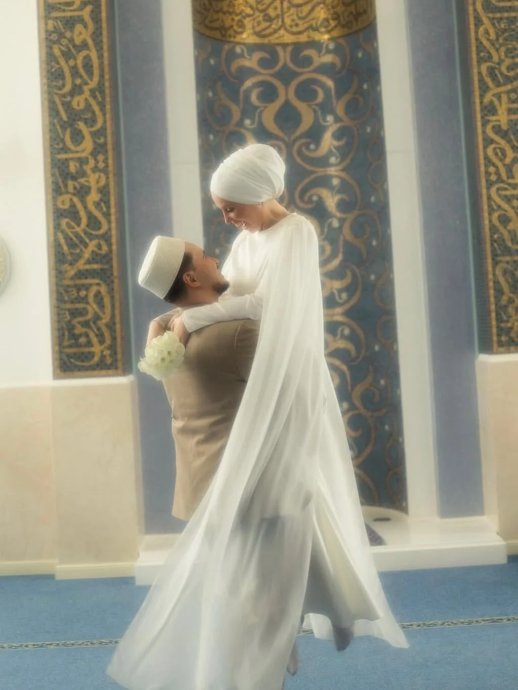 A bride and groom in traditional Islamic wedding attire, with the groom lifting the bride inside a mosque, featuring blue and gold Islamic calligraphy and patterns in the background.