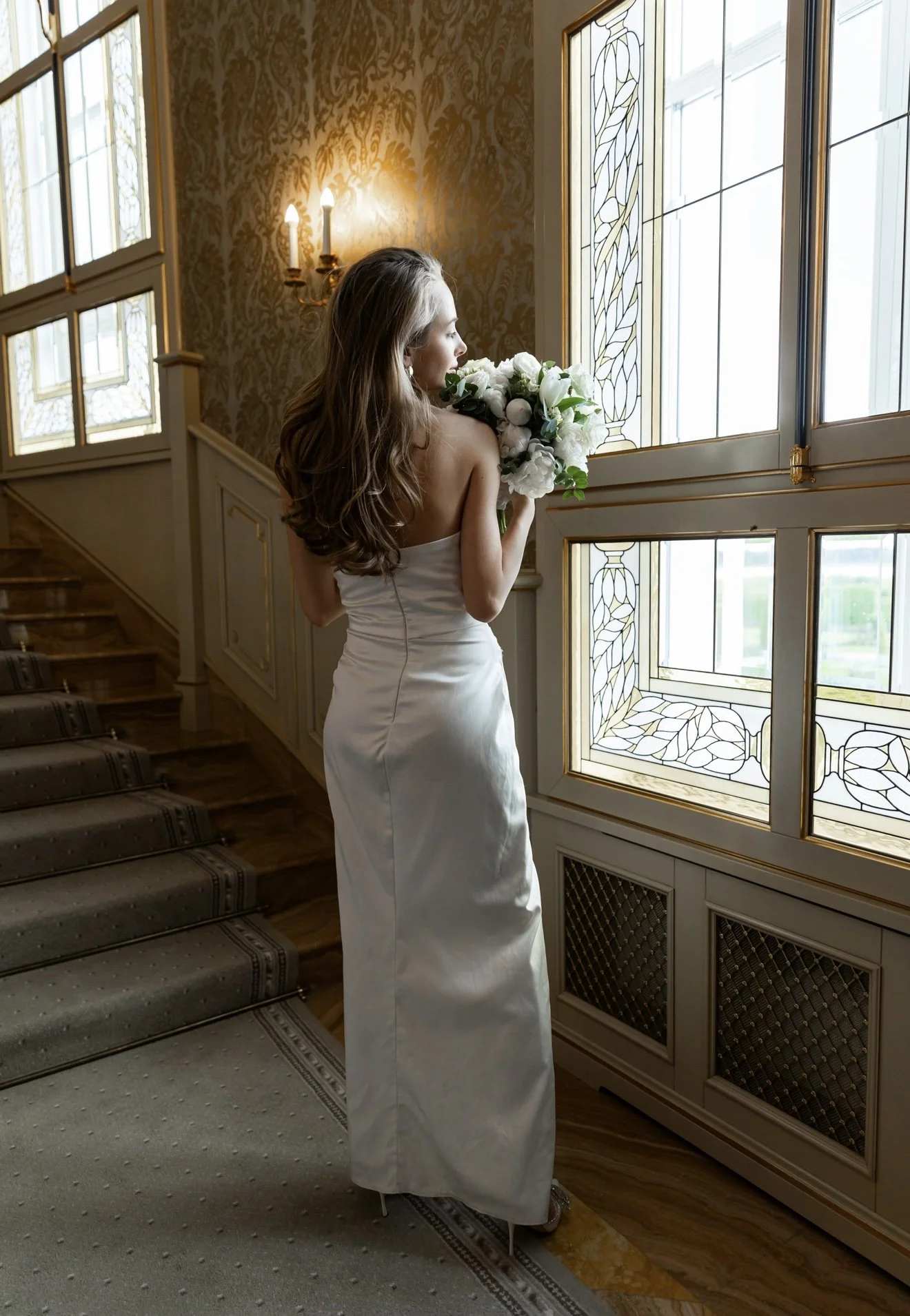A woman in a white dress holding a bouquet of white flowers, standing near a window with stained glass, in an elegant interior with patterned wallpaper and a stairway with a patterned carpet.