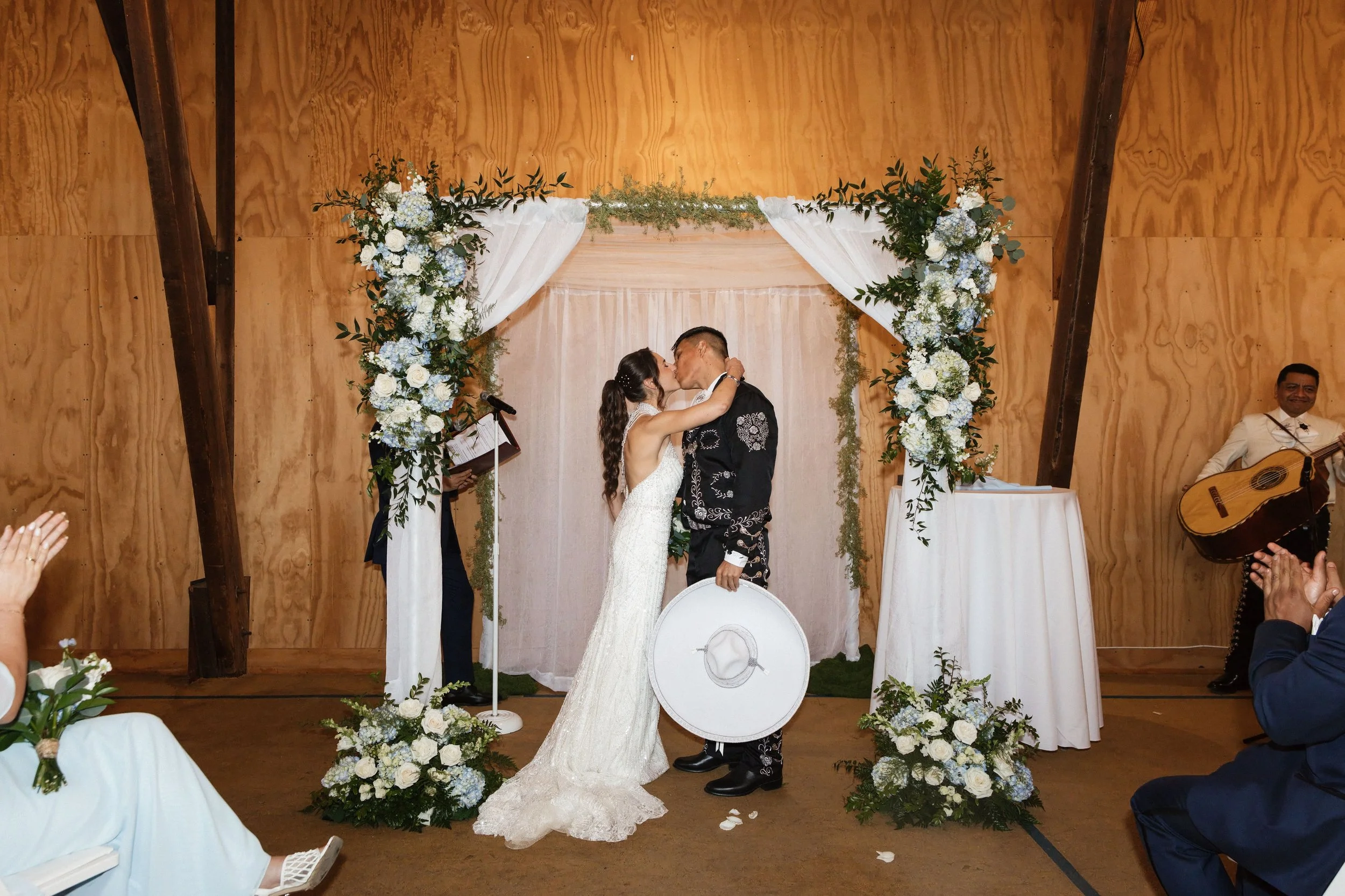 A wedding ceremony with a bride and groom sharing a kiss at an altar decorated with white and light blue flowers and greenery, indoors with wooden walls and beams, musicians are visible on the right side, and guests are seated watching the couple.