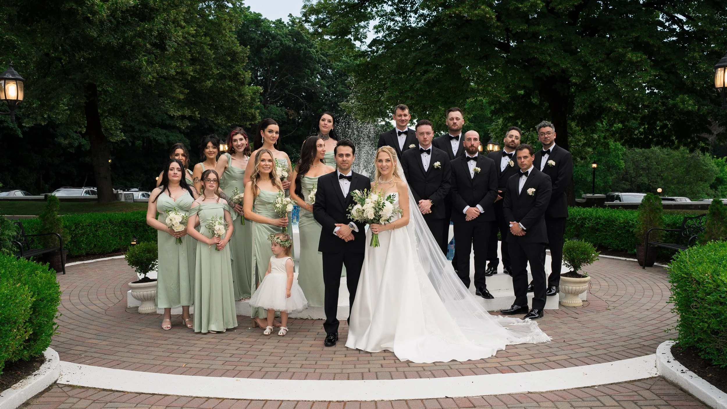 A large group of people dressed in formal attire, including wedding clothes, standing outdoors near a fountain with green trees in the background. The group includes brides, grooms, bridesmaids, groomsmen, and children.