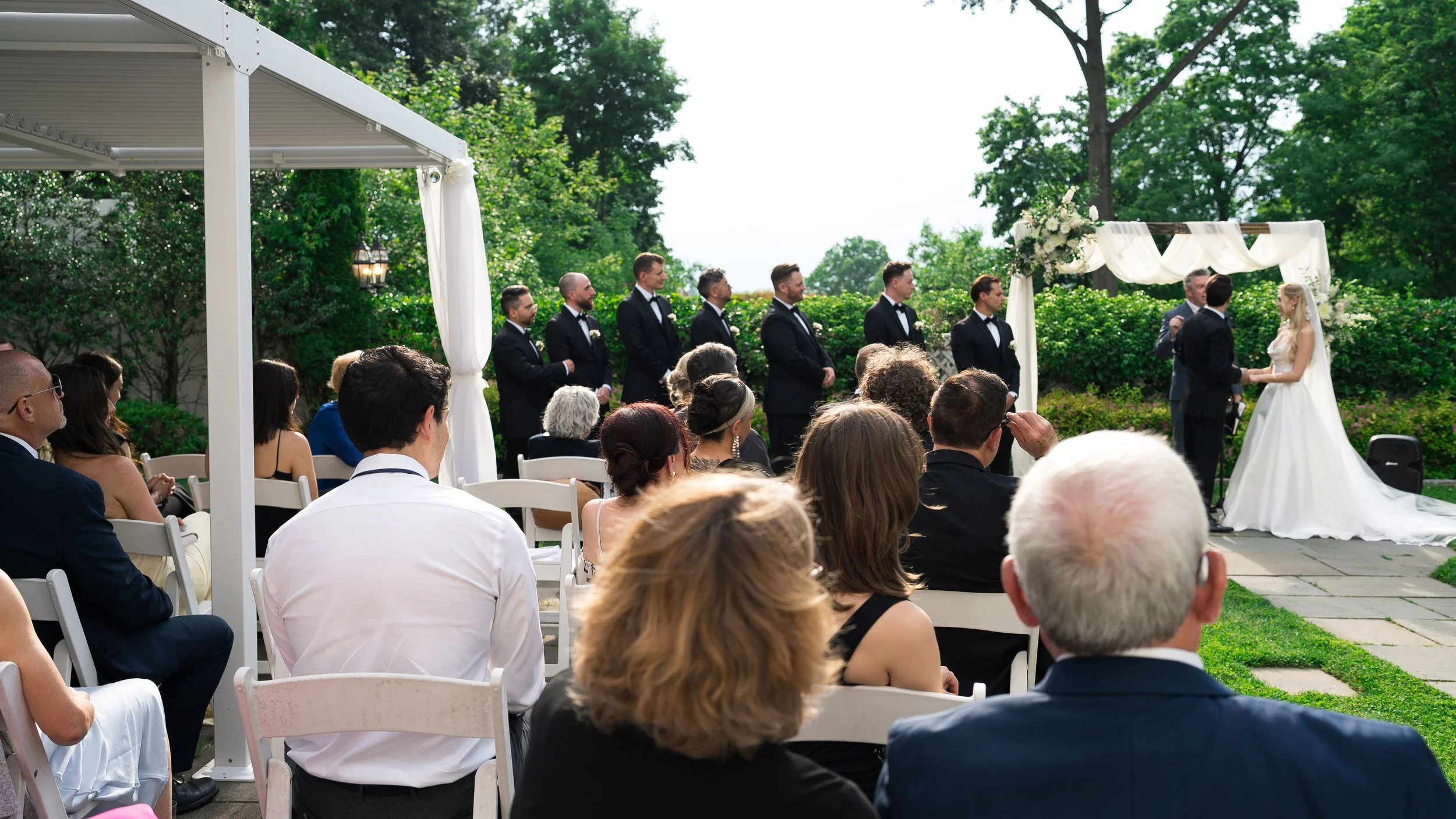 Outdoor wedding ceremony with bride and groom exchanging vows, surrounded by guests seated under a white canopy, lush green trees in the background.