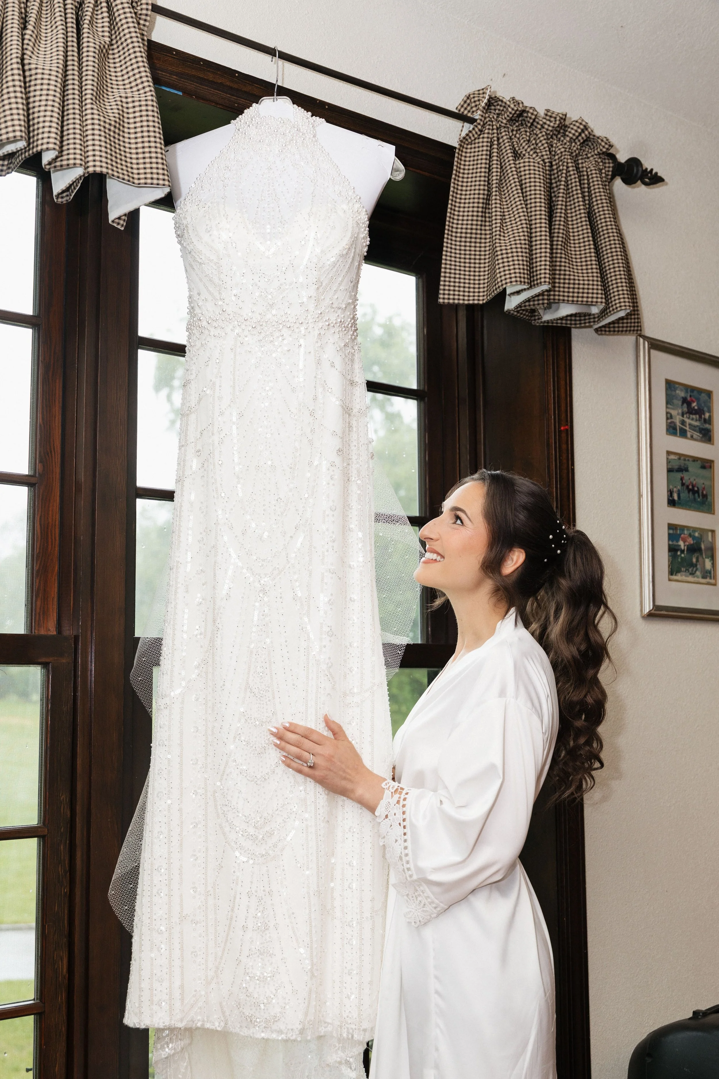 A woman in a white satin robe is smiling and looking at a wedding dress hanging in front of a window. She has long wavy brown hair with pearl hairpins.