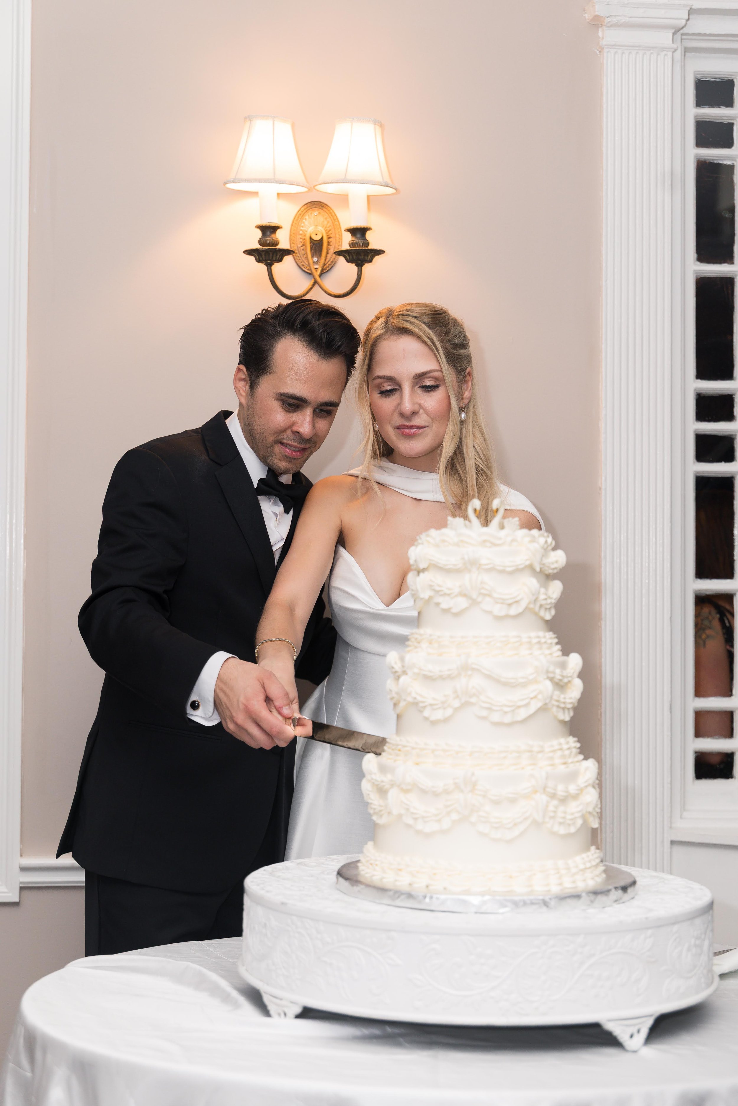 A newlywed couple in formal attire cutting a wedding cake together at their wedding reception. The groom is wearing a black tuxedo with a bow tie, and the bride is wearing a white wedding dress. The wedding cake has multiple tiers decorated with crea