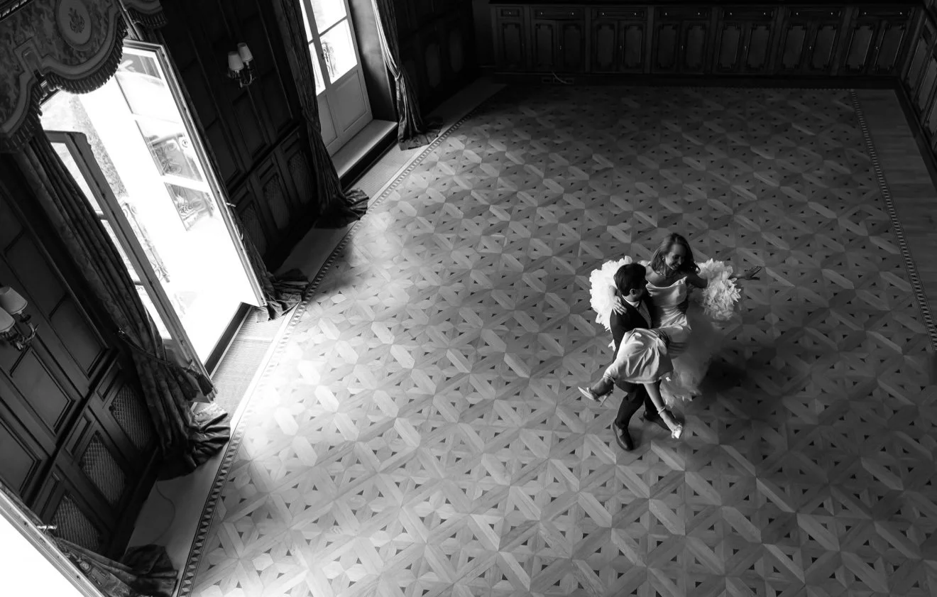 A top-down black and white photo of a woman in a wedding dress dancing with two children on a patterned wooden floor in a room with large windows, dark wood paneling, and curtains.
