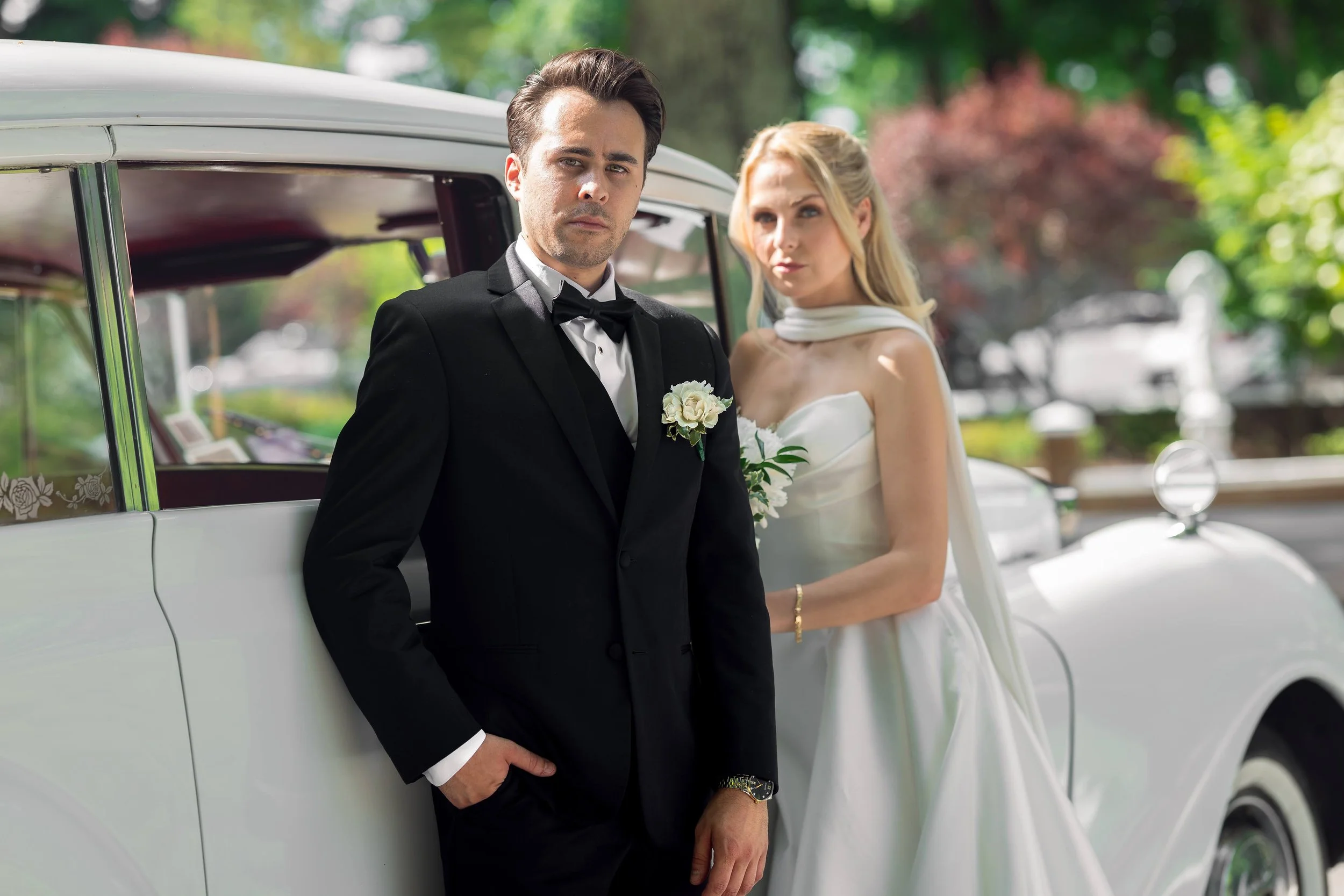 A man in a tuxedo and a woman in a wedding dress standing next to a vintage white car outdoors.