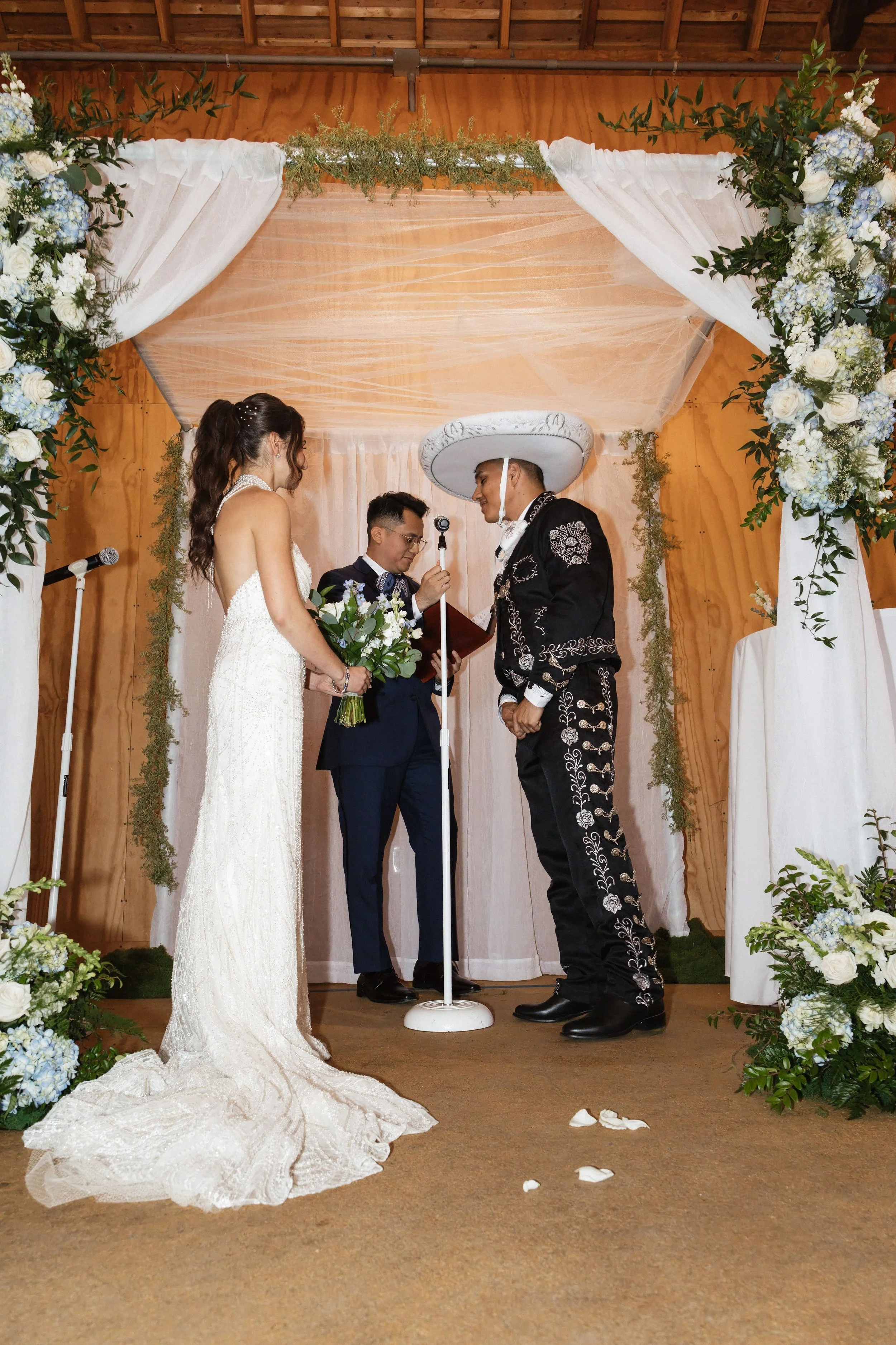 A wedding ceremony with a bride and groom standing under an arch decorated with white and blue flowers, white drapes, and greenery, while the officiant reads from a book.