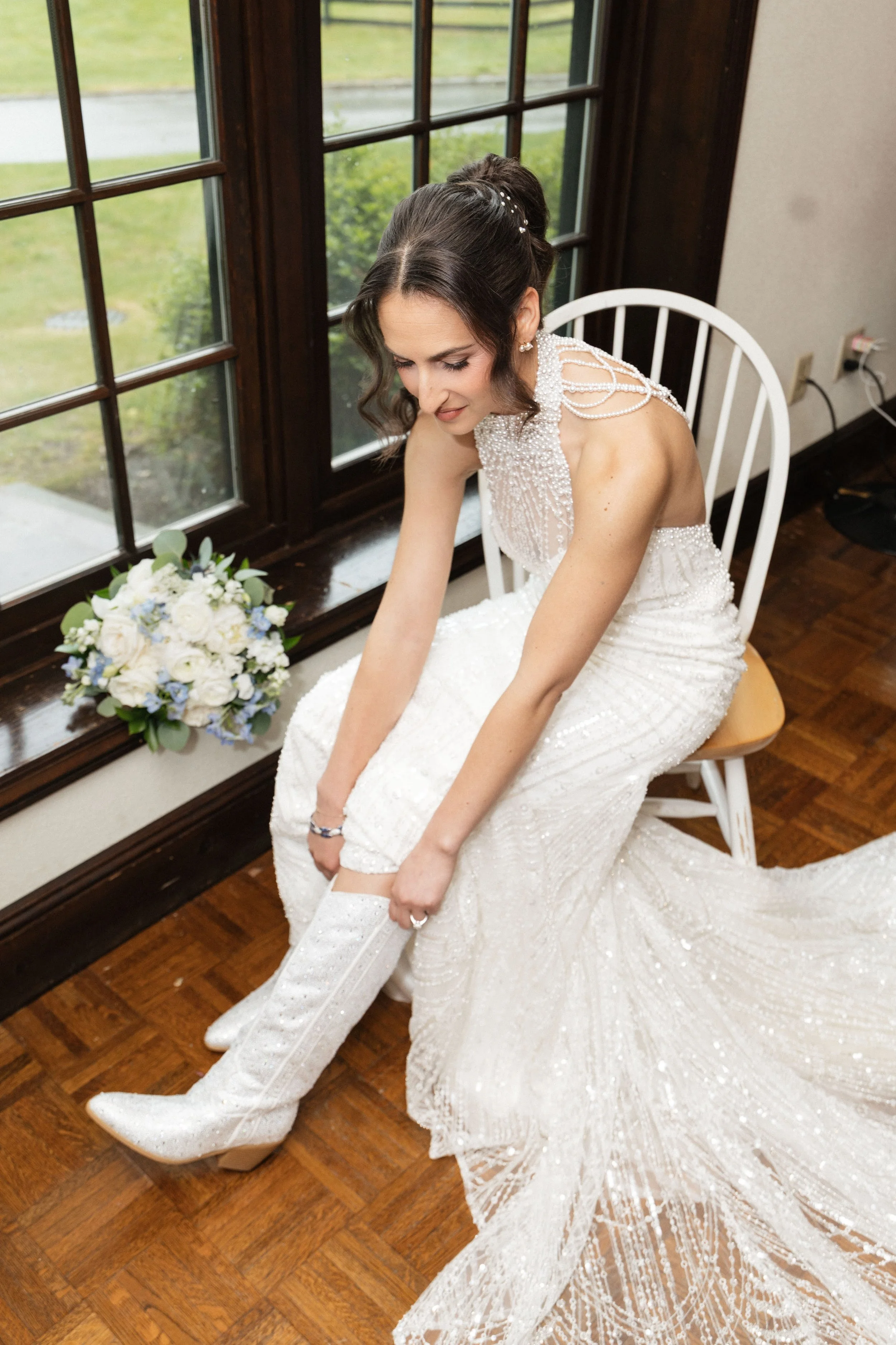 A bride sitting on a wooden chair in front of a window, adjusting her thigh-high white stocking, wearing a pearl-embellished wedding dress, with a bouquet of white and blue flowers nearby.