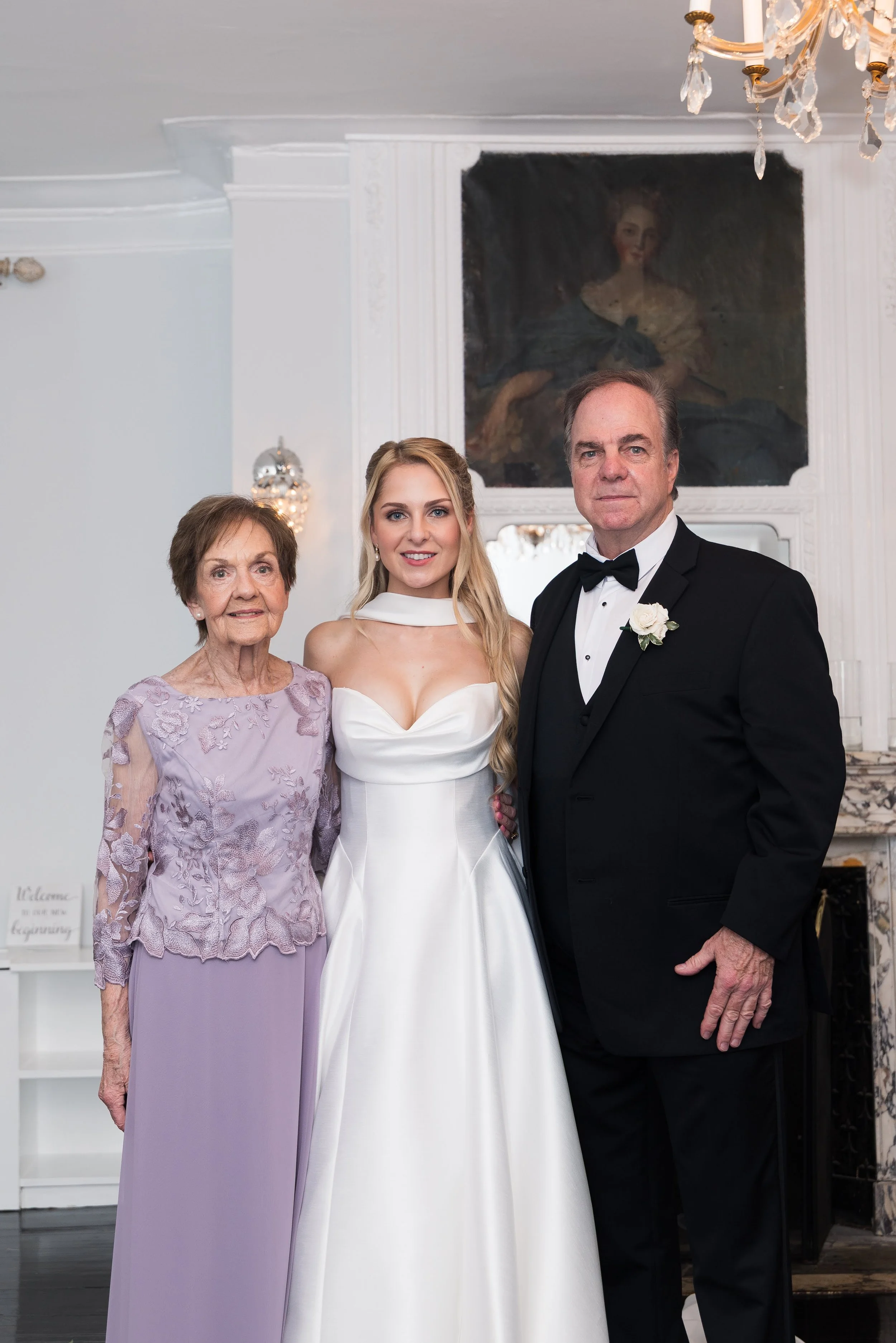 A wedding scene featuring three people: an elderly woman in a lavender dress with lace embroidery, a young woman in a white wedding gown, and a middle-aged man in a black tuxedo with a white boutonniere. They are standing inside a well-decorated room