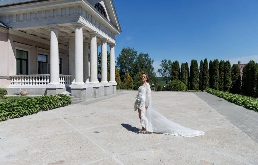 Woman in a white wedding dress standing on a large, open driveway in front of a grand, classical-style mansion with white columns and a balcony, with green trees and a blue sky in the background.