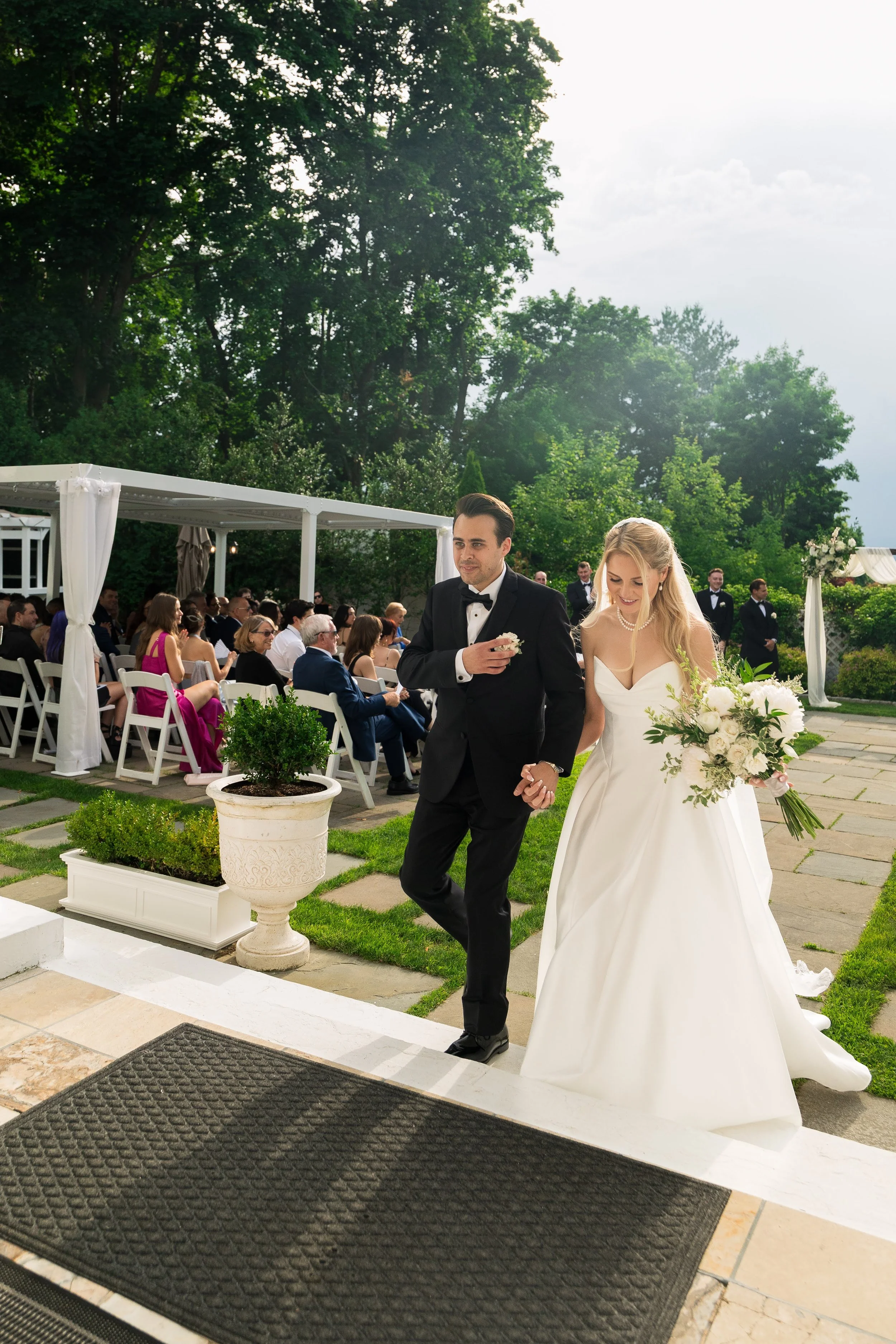 A bride and groom walking hand in hand at their outdoor wedding ceremony. The bride is holding a large bouquet of white flowers, and the groom is dressed in a black tuxedo with a bow tie. Guests are seated and watching in the background under a white