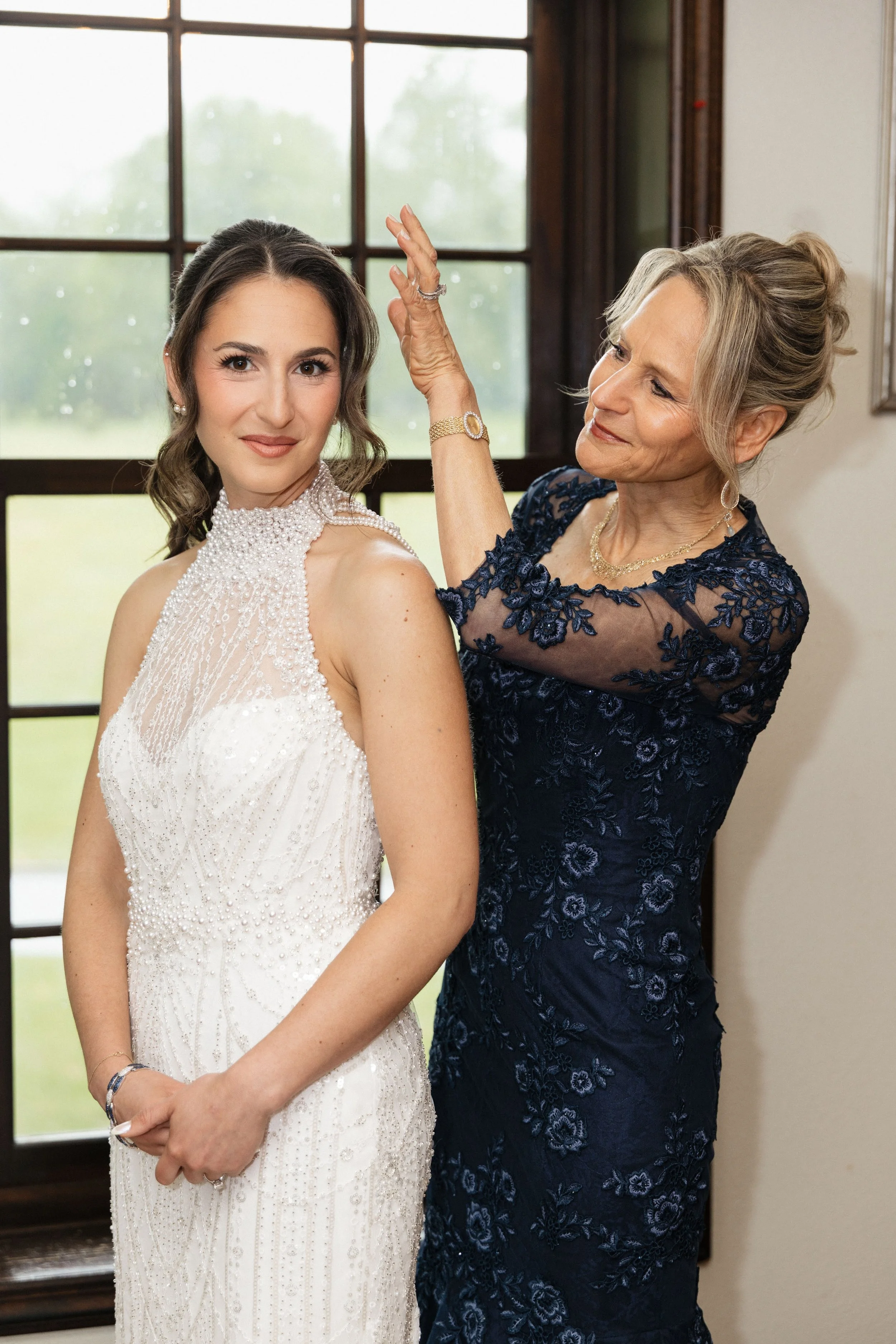 A young woman in a white wedding dress with pearl embellishments is standing indoors, while an older woman in a navy blue floral dress is adjusting her hair and smiling.