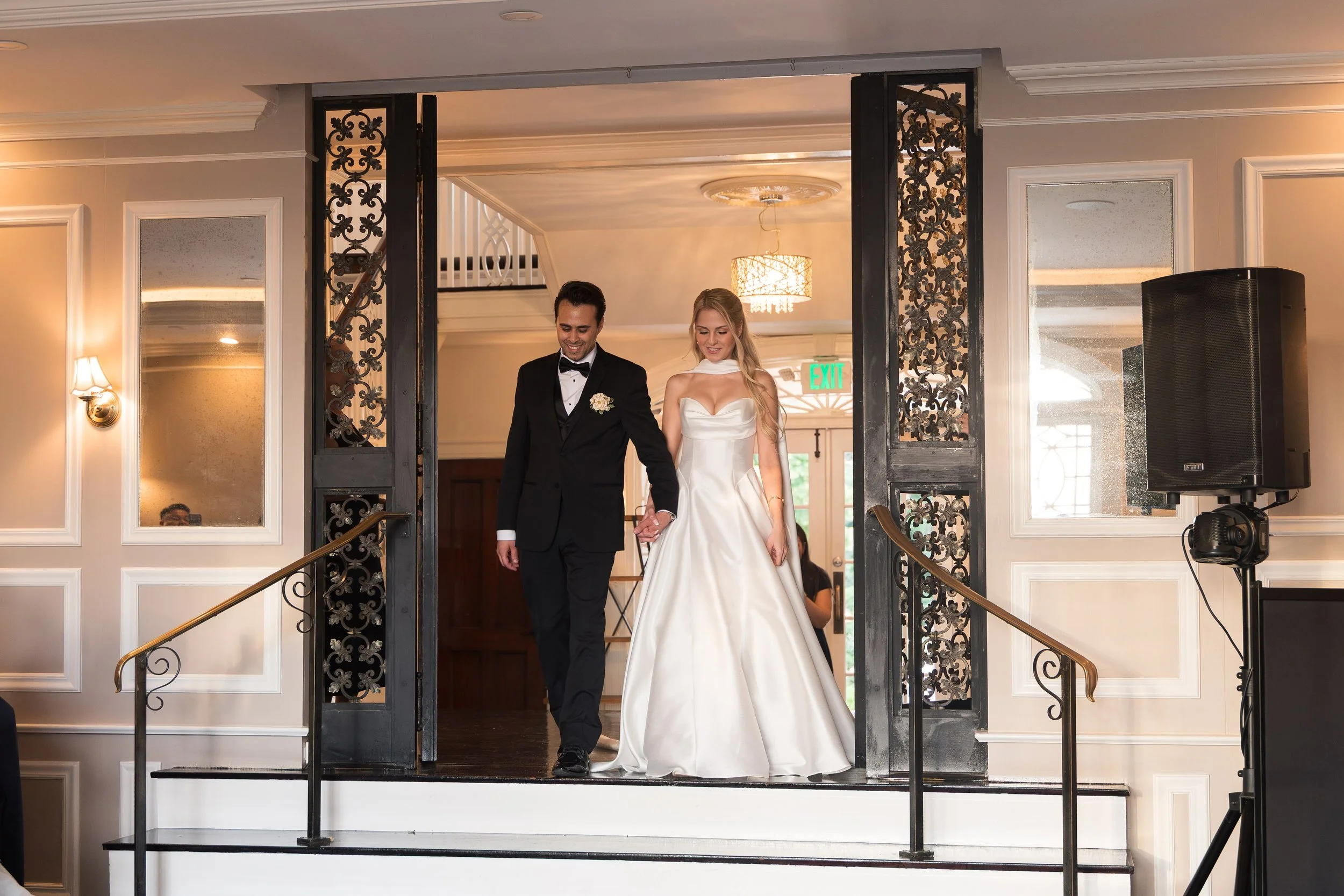 A bride and groom walking down the stairs at their wedding ceremony. The bride wears a white dress and the groom wears a black tuxedo. They are holding hands and smiling.