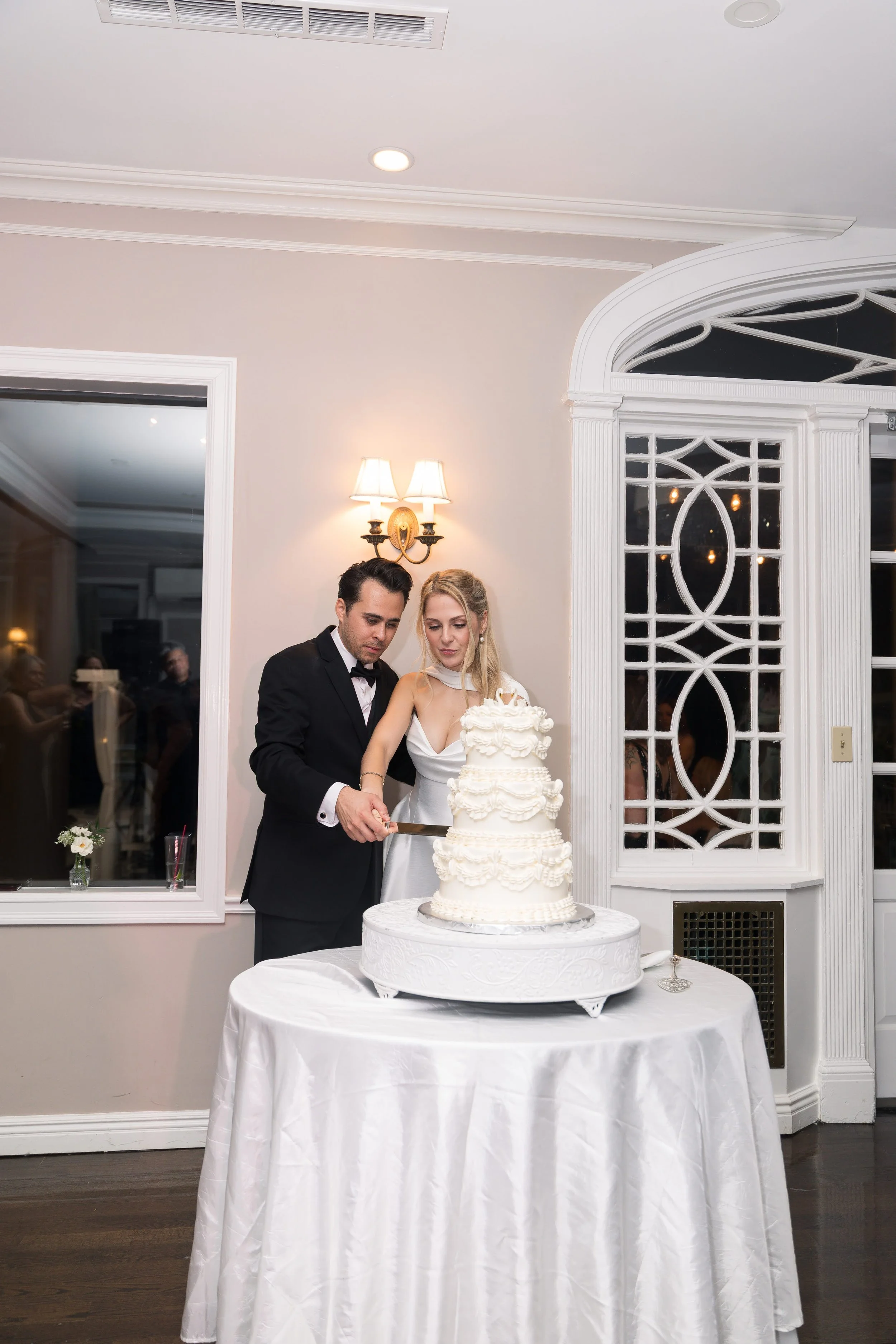 A bride and groom cut a wedding cake together at their wedding reception. The bride wears a white gown and the groom wears a black tuxedo. The wedding cake is large, white, and decorated with multiple layers and intricate designs. The reception area 