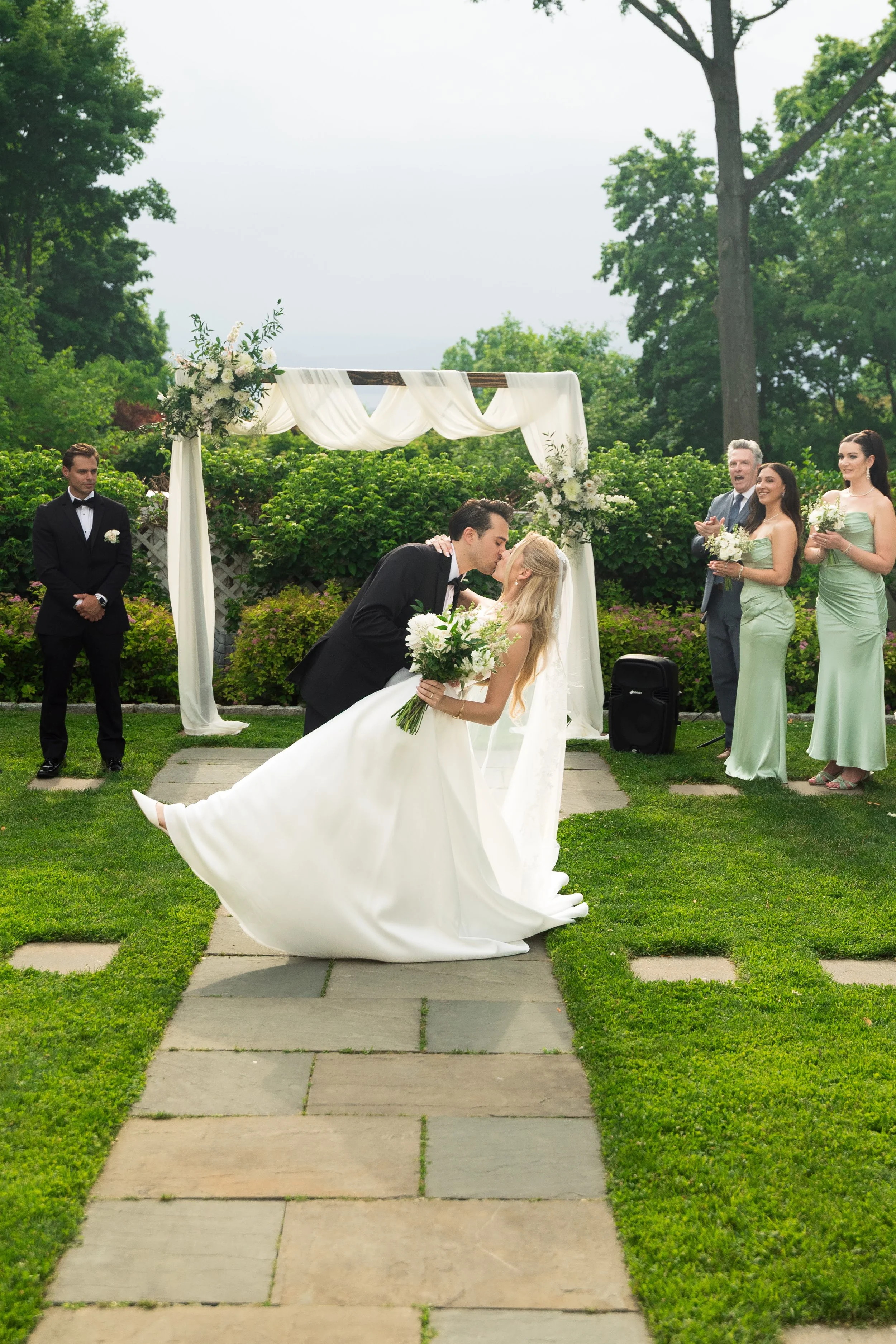 A bride and groom sharing a kiss under a wedding arch during an outdoor wedding ceremony, with the bride holding a bouquet and wedding party members in the background.