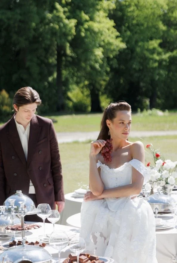 A woman in a white wedding dress and a man in a dark suit are outdoors near a table with glassware and desserts. The woman holds a bunch of grapes at her neck, looking to her right, while the man looks down.