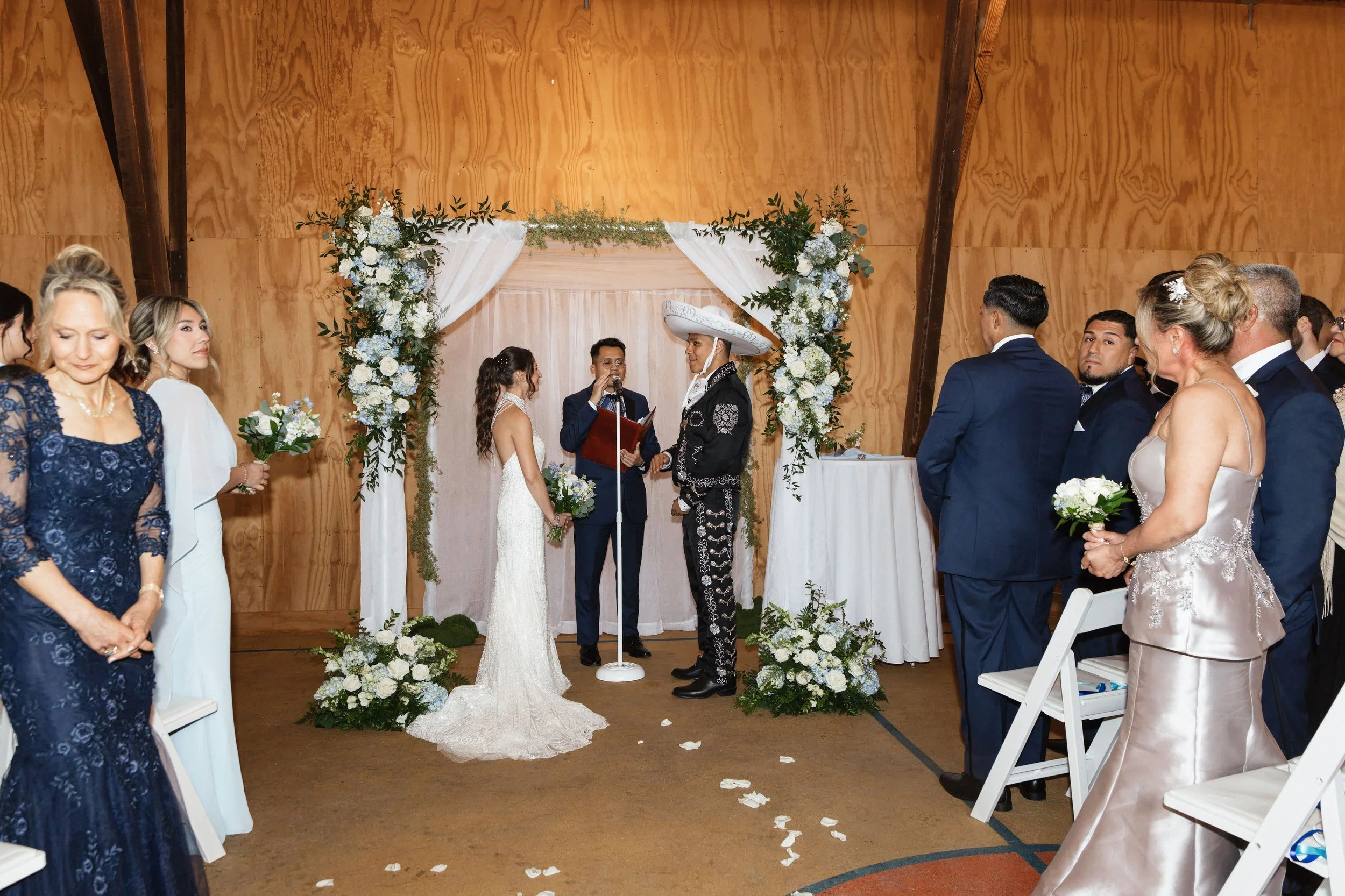 A wedding ceremony with a couple exchanging vows under a floral arch, surrounded by seated guests in formal attire.