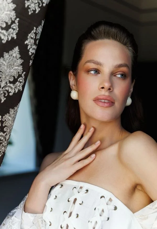 A young woman with fair skin, blue eyes, and brown hair with blonde highlights, styled back. She wears large, round white earrings and a white strapless top with a lace-up front. She gazes slightly to the side with a contemplative expression, touchin