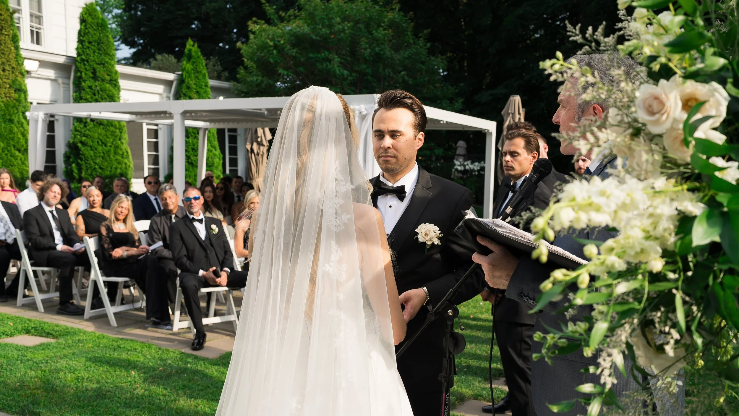 Bride and groom exchanging vows at an outdoor wedding ceremony, with guests seated in the background and officiant holding a microphone and book.